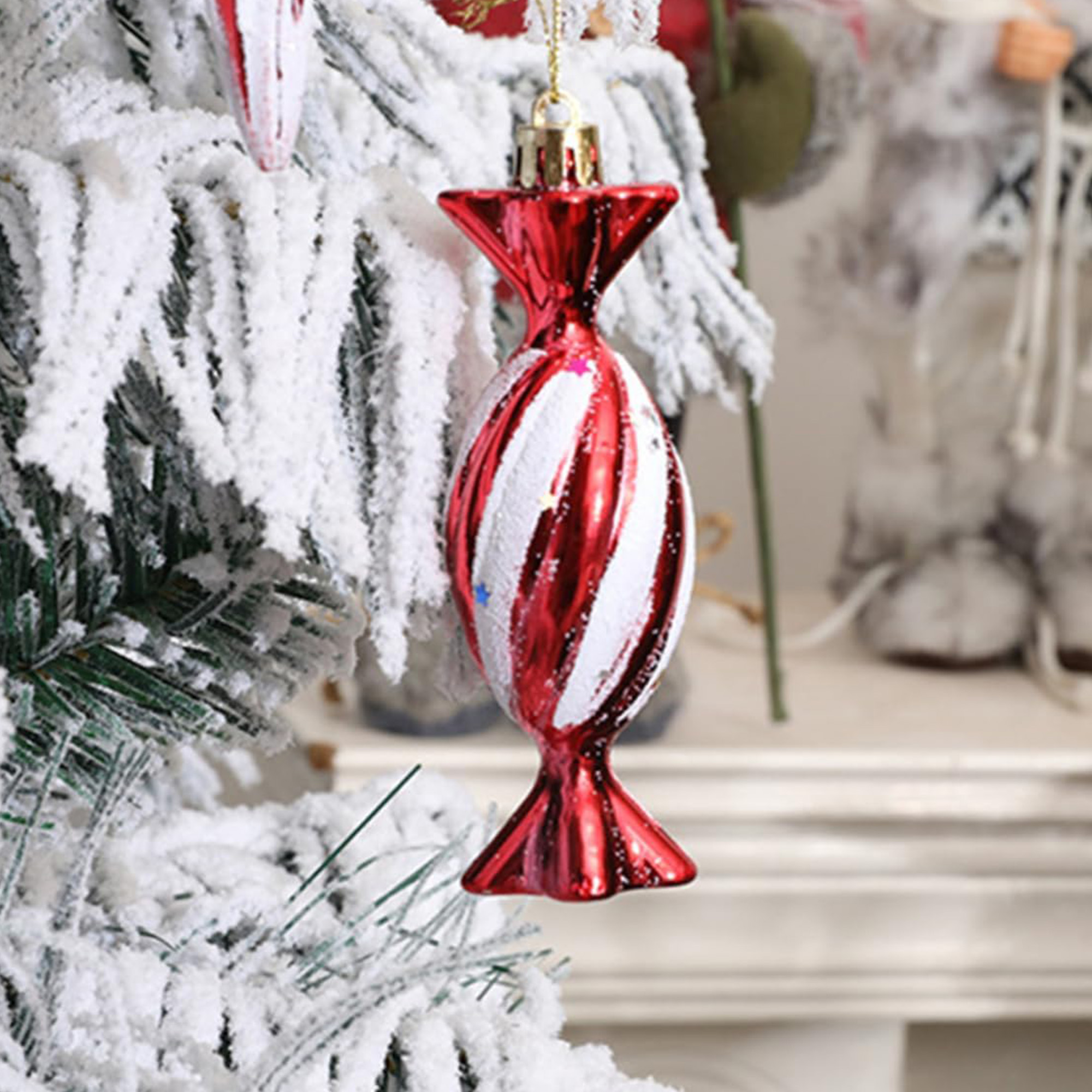 Un adorno navideño rojo y blanco con forma de caramelo cuelga de un árbol nevado, con un fondo festivo borroso.
