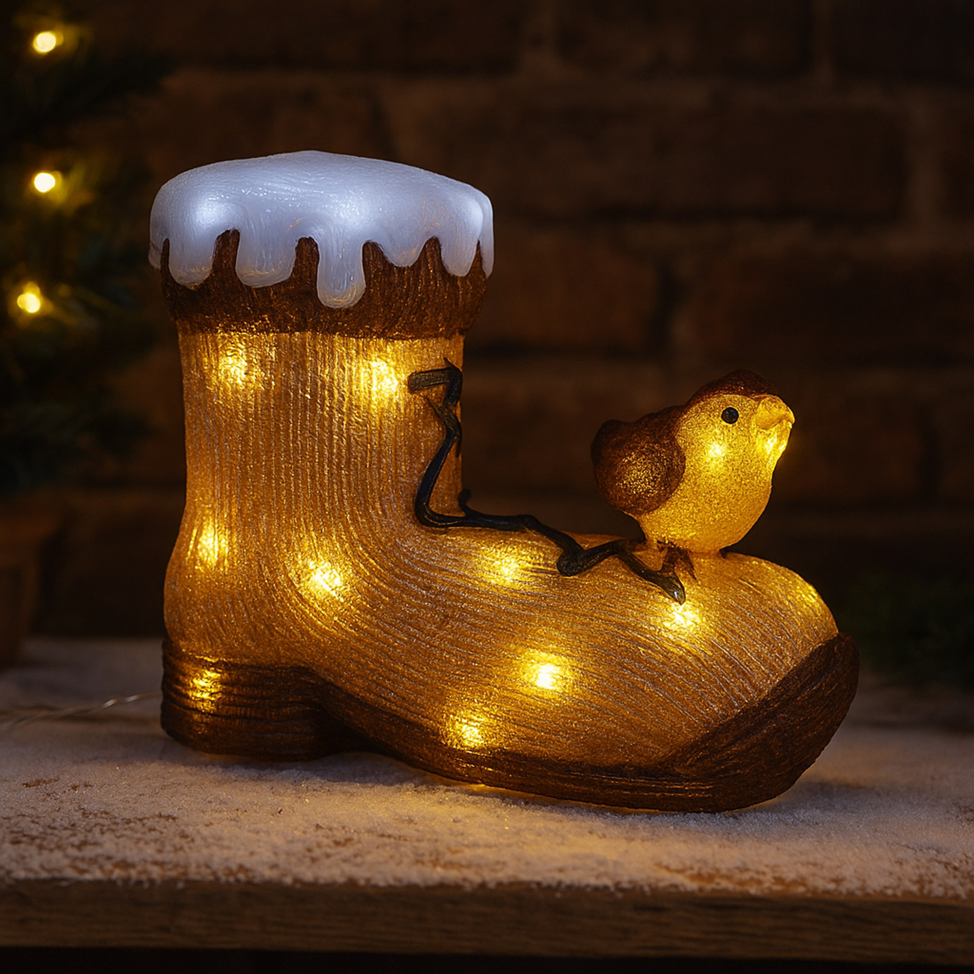 Una bota decorativa iluminada con un pájaro encima, colocada sobre una superficie nevada contra una pared de ladrillos.
