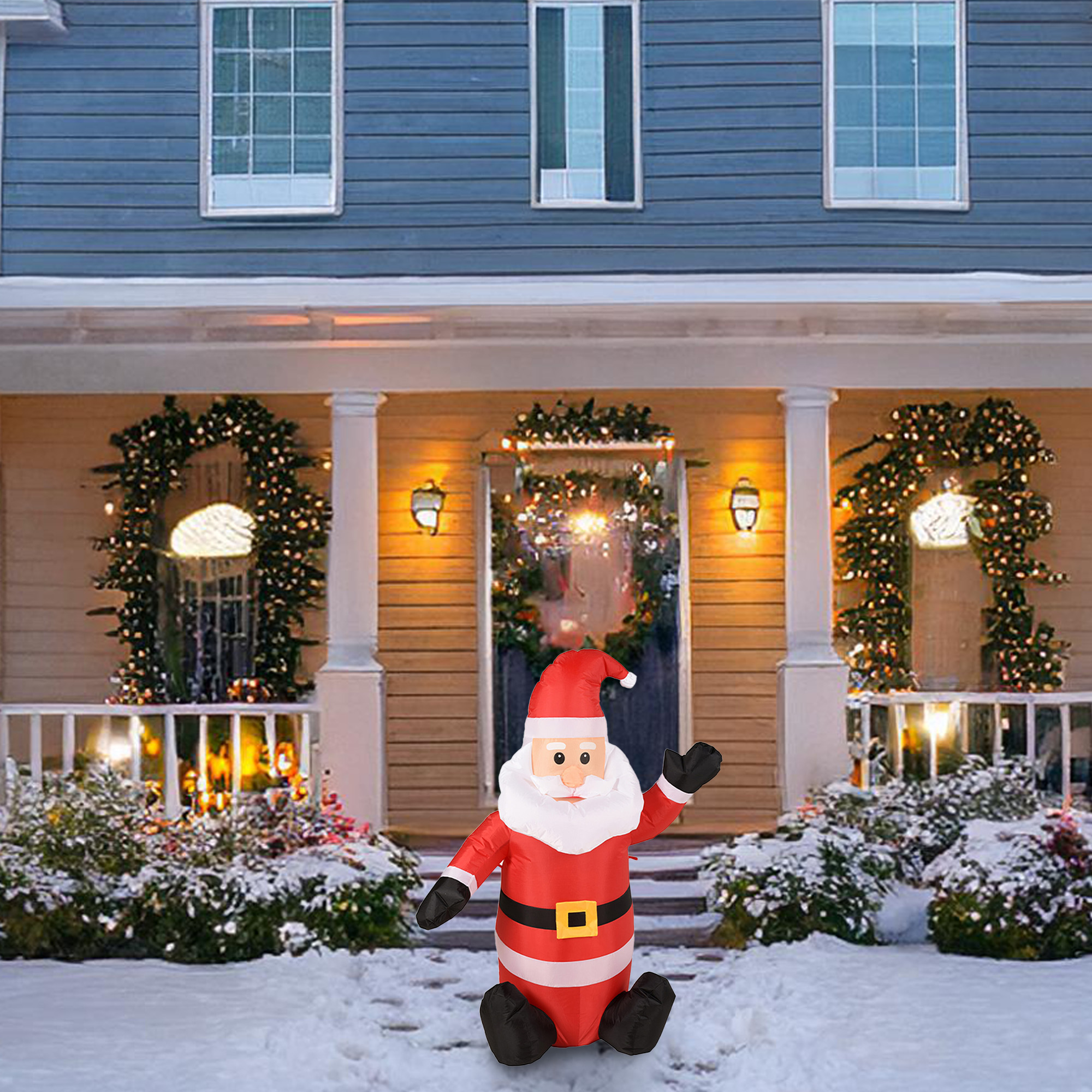 Un Papá Noel inflable se sienta en la nieve frente a una casa decorada para Navidad. La casa tiene coronas y luces.