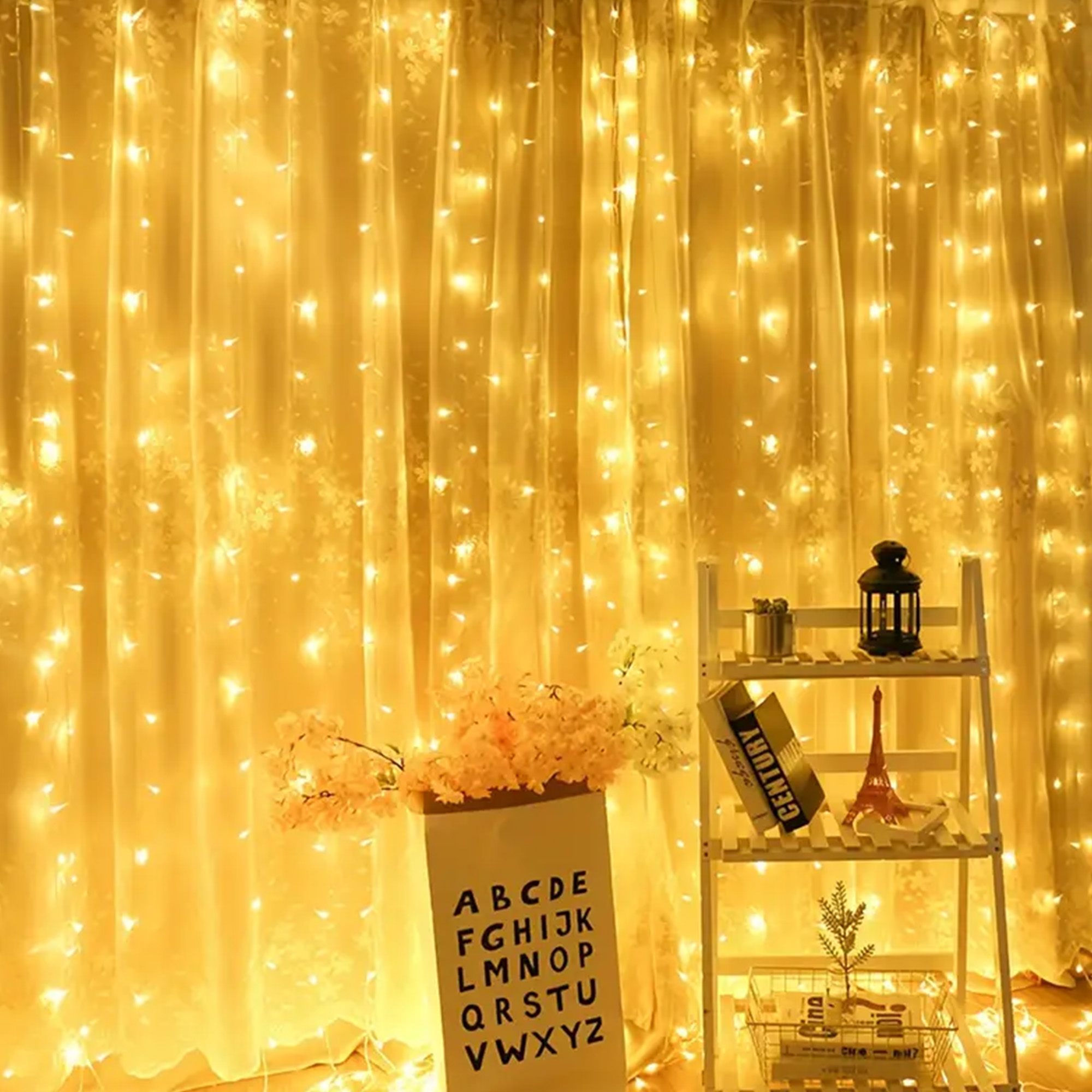 Cortina amarilla iluminada con luces de hadas. Flores en una bolsa de papel con alfabeto. Una estantería con decoraciones a la derecha.