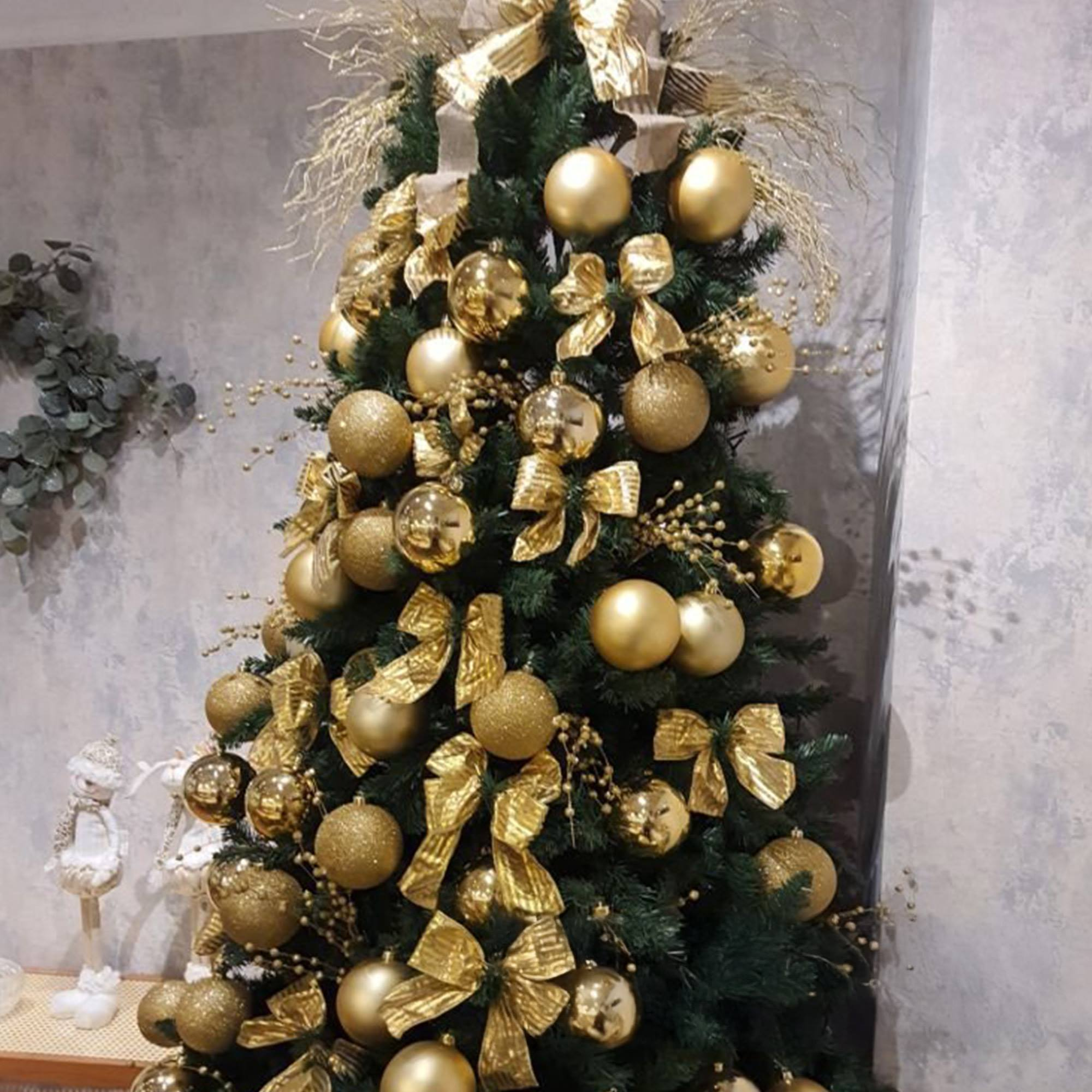 Un árbol de Navidad decorado con adornos y lazos dorados se alza contra una pared gris con un muñeco de nieve.