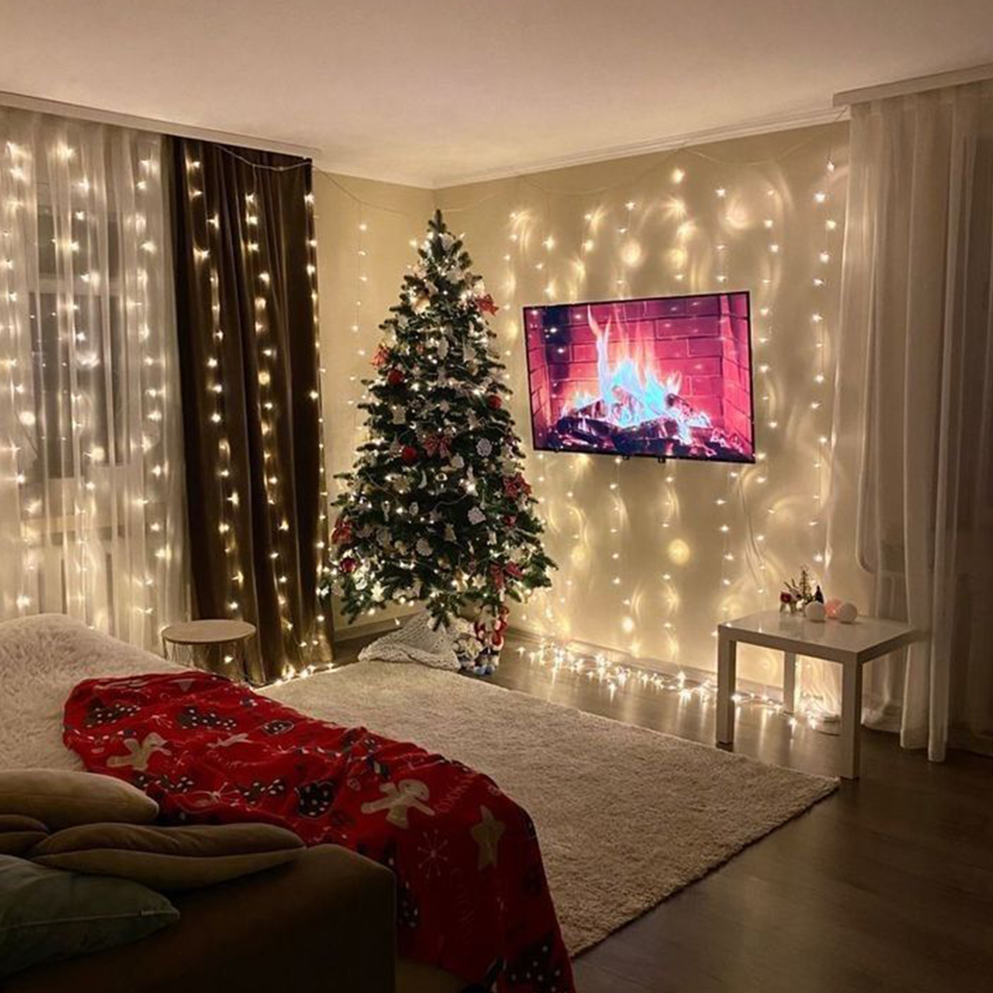Un árbol de Navidad decorado en una habitación acogedora con luces. Un televisor muestra una escena de chimenea. Manta roja en el sofá.