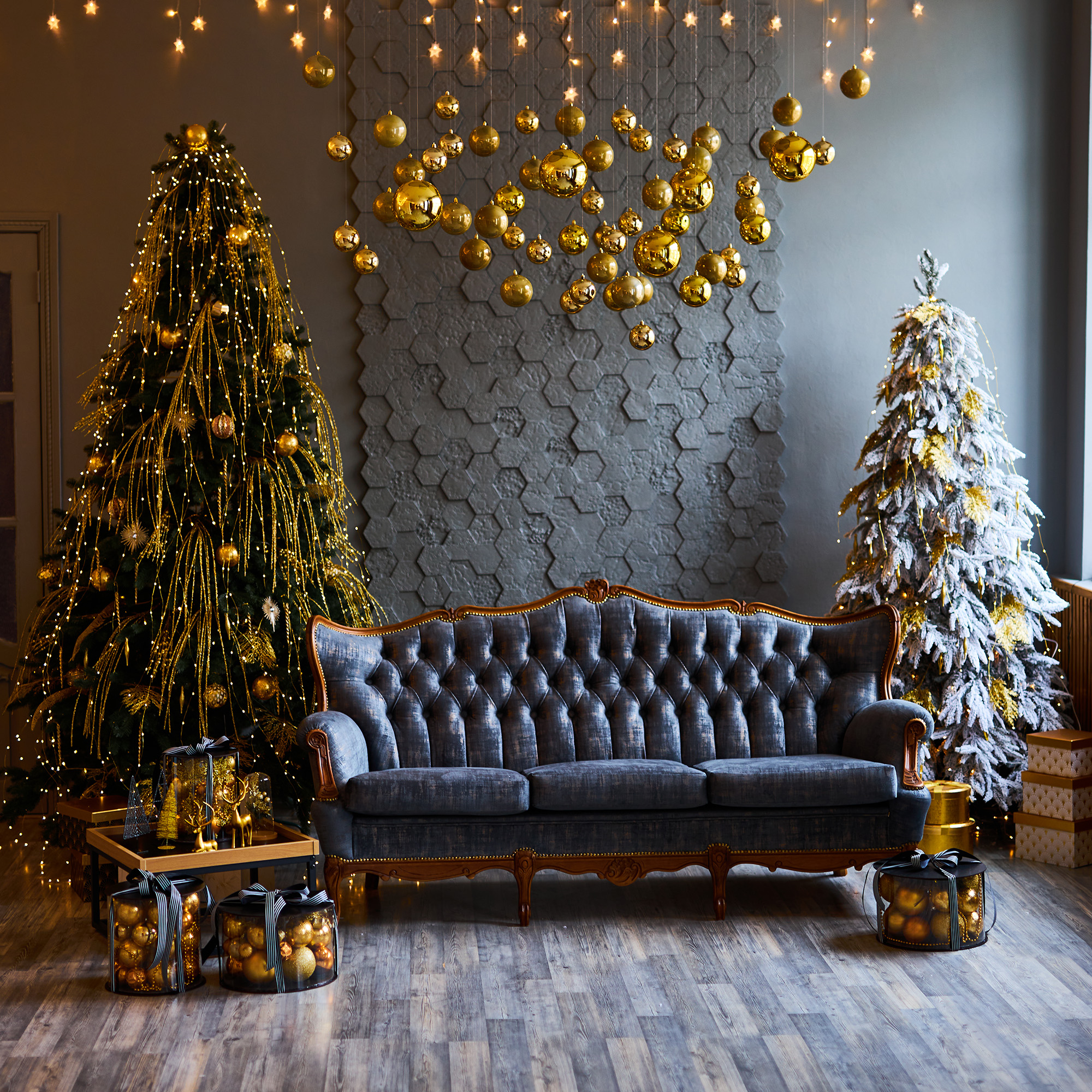 Una habitación decorada con dos árboles de Navidad, un sofá y adornos dorados colgantes. Los regalos y las luces añaden ambiente festivo.