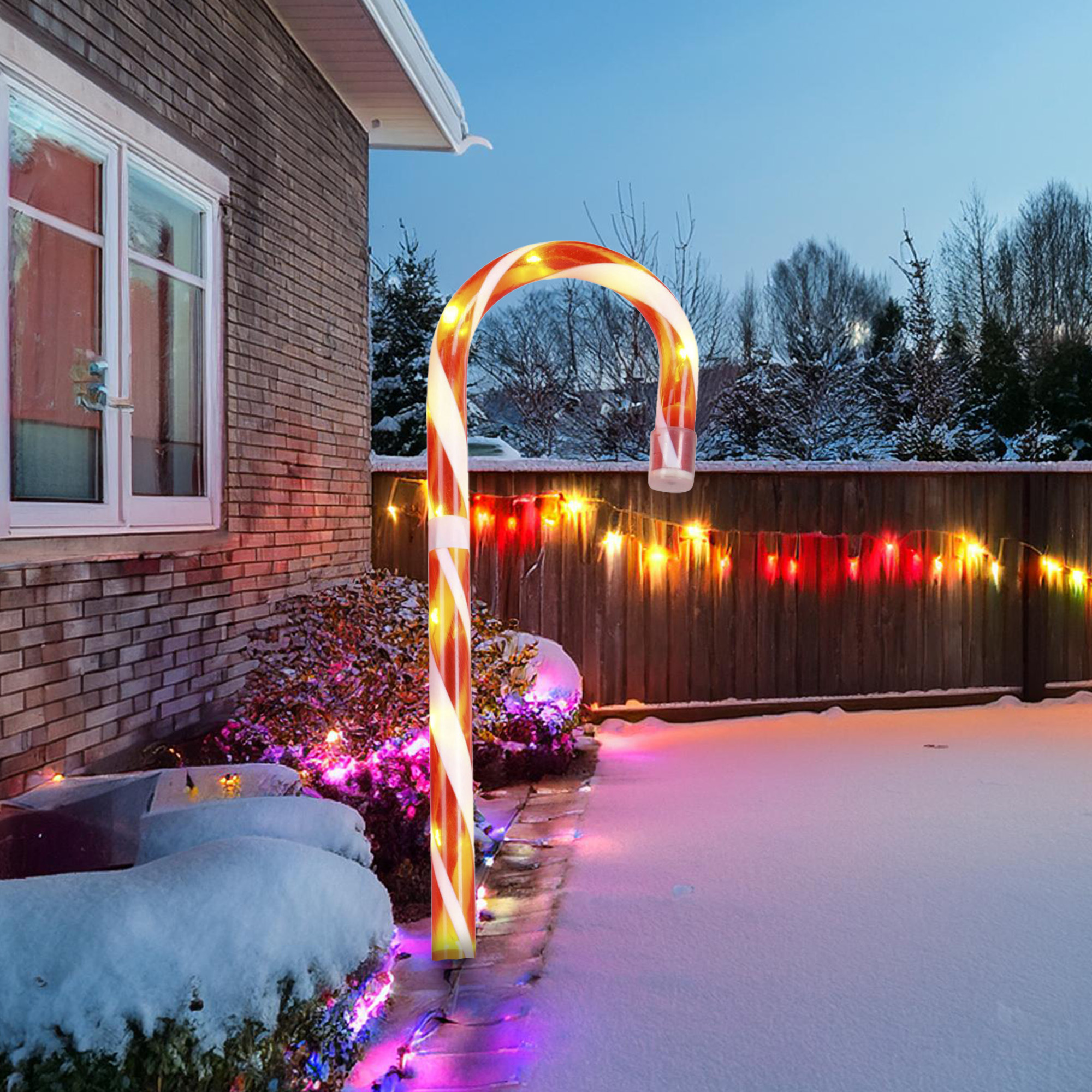 Decoración de bastón de caramelo iluminado en un jardín nevado, cerca de una casa y una valla, al anochecer.