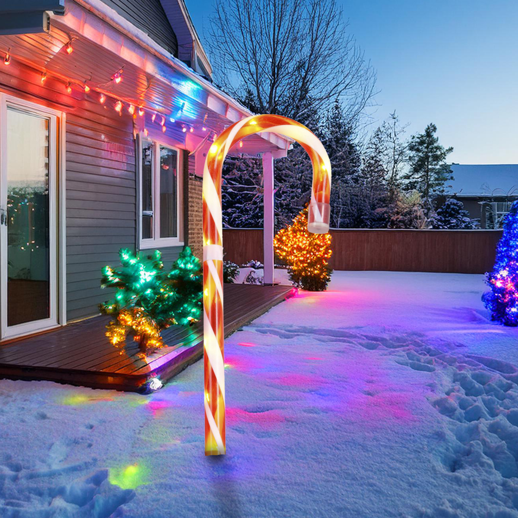Una decoración gigante de bastón de caramelo en un jardín nevado, con luces navideñas en la casa y los árboles. Escena nocturna.