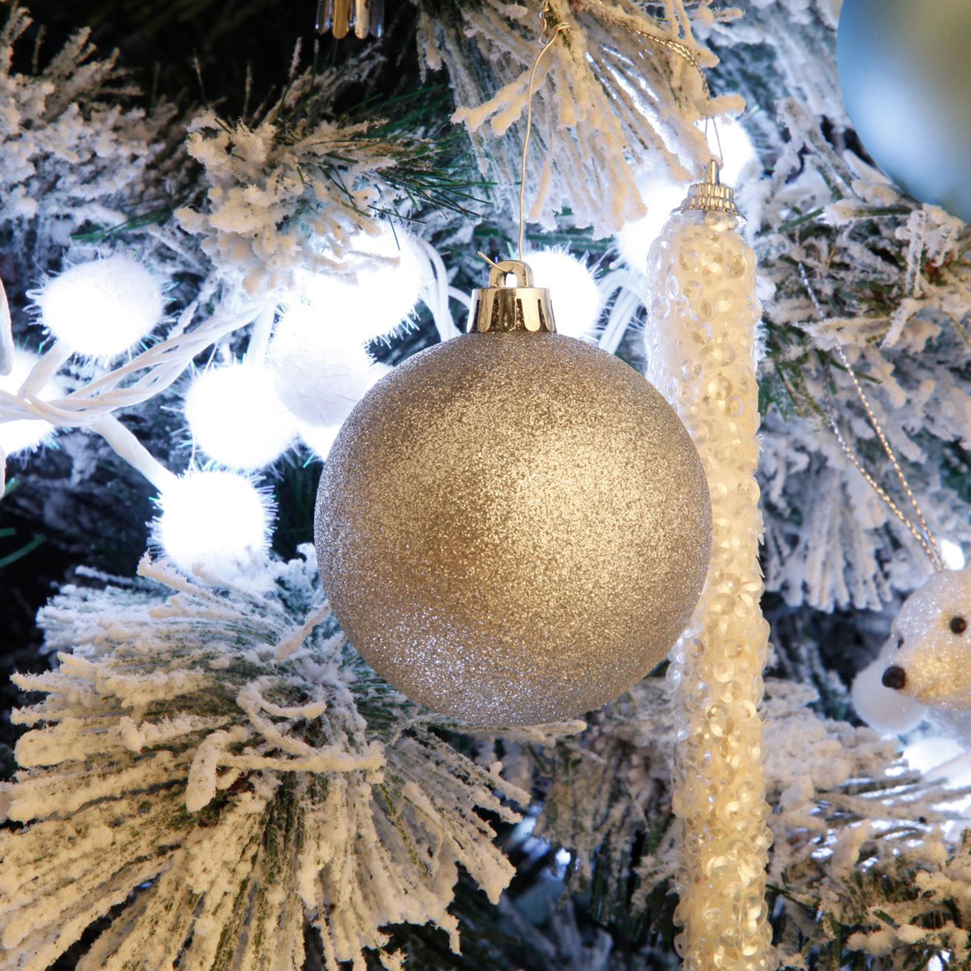 Un árbol de Navidad con un adorno plateado enfocado. Luces blancas y ramas heladas rodean la bola.