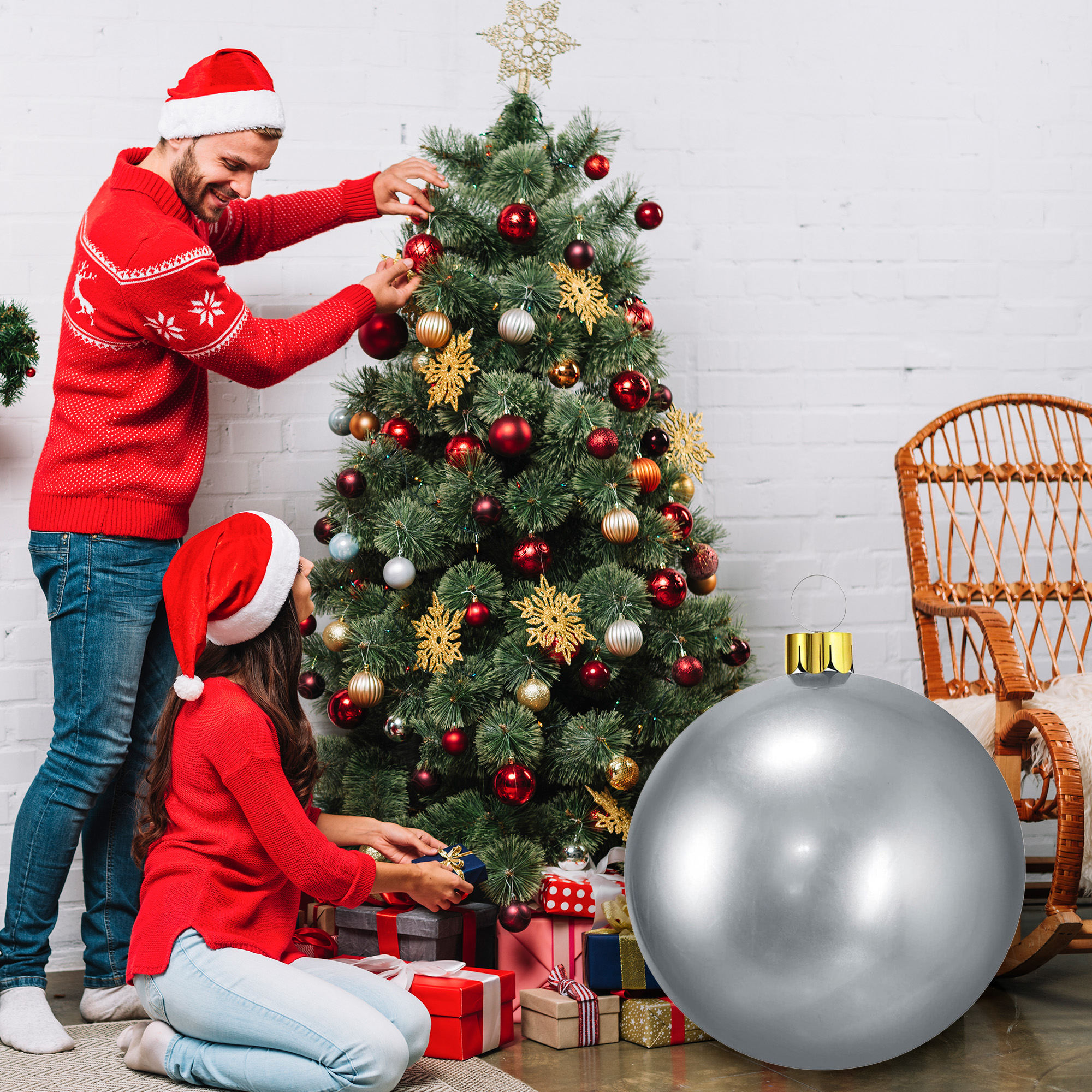 Pareja decora árbol de Navidad. Hombre en suéter rojo, mujer con gorro de Papá Noel, rodeados de regalos y adorno plateado.