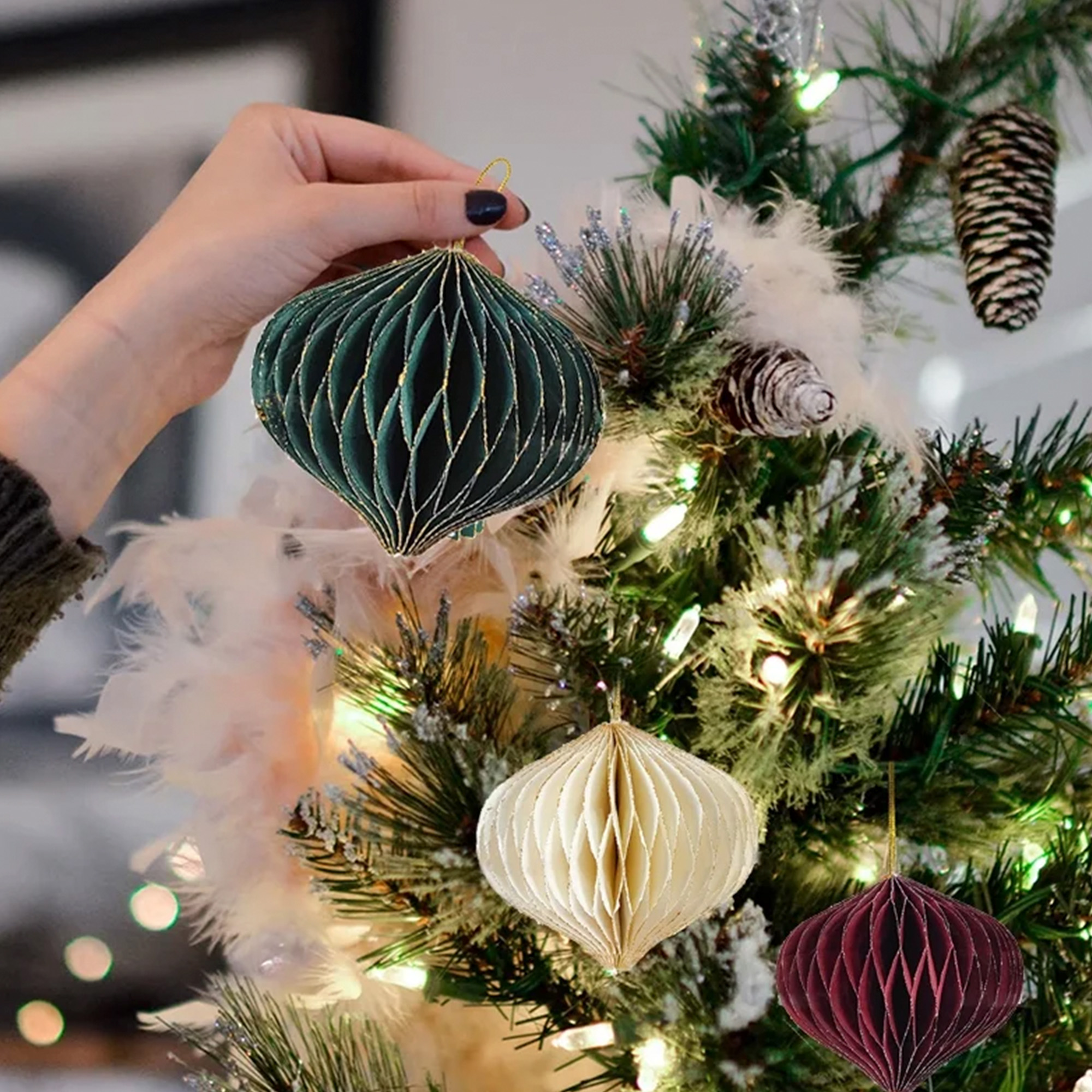 Una mano sostiene un adorno de papel verde en un árbol de Navidad con otros adornos y luces.