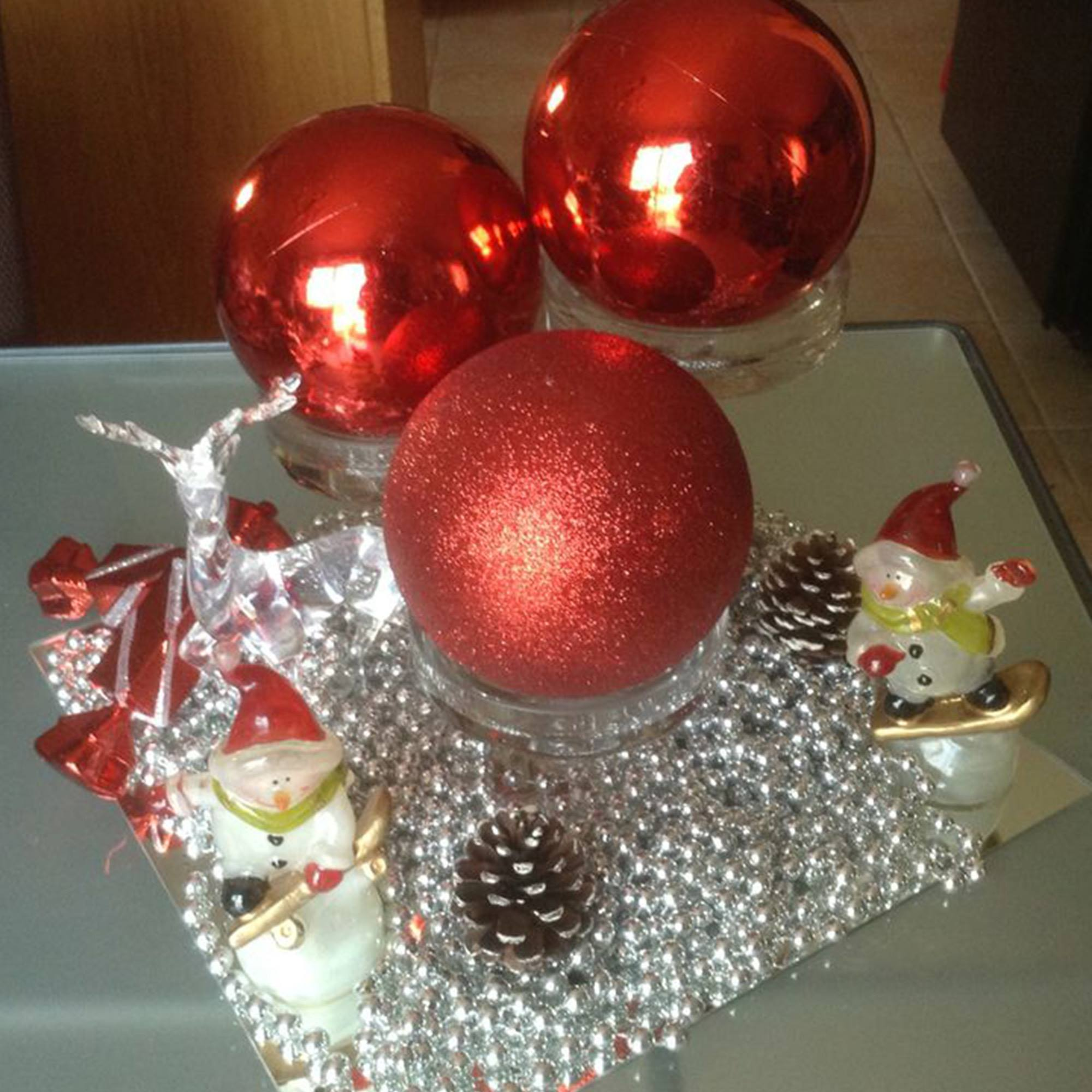 Decoraciones navideñas: adornos rojos, figuras de muñecos de nieve, piñas y cuentas plateadas sobre una superficie reflectante.