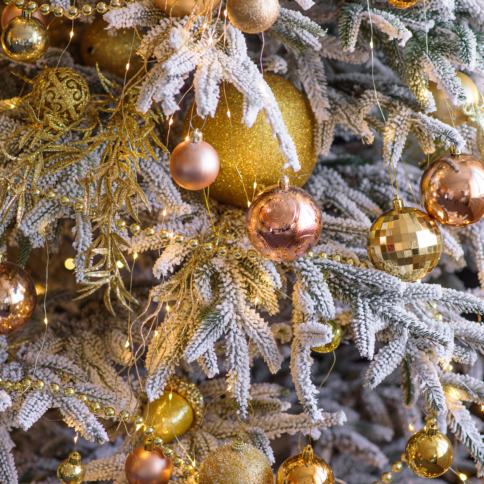 Un árbol de Navidad decorado con adornos dorados, cuentas y luces. Las ramas están cubiertas de nieve artificial.