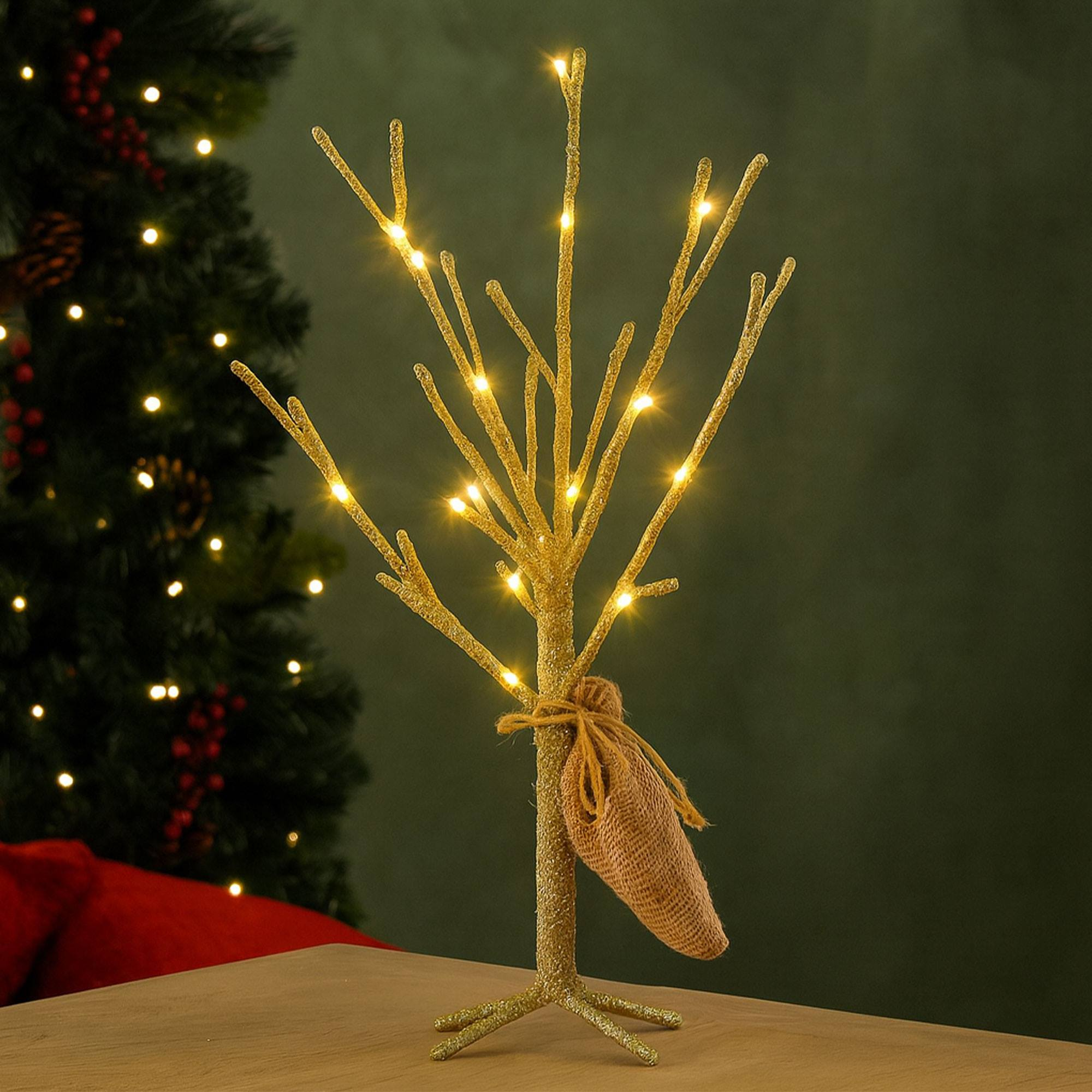 Un árbol decorativo dorado con luces y un saco de arpillera atado. Un árbol de Navidad en el fondo.