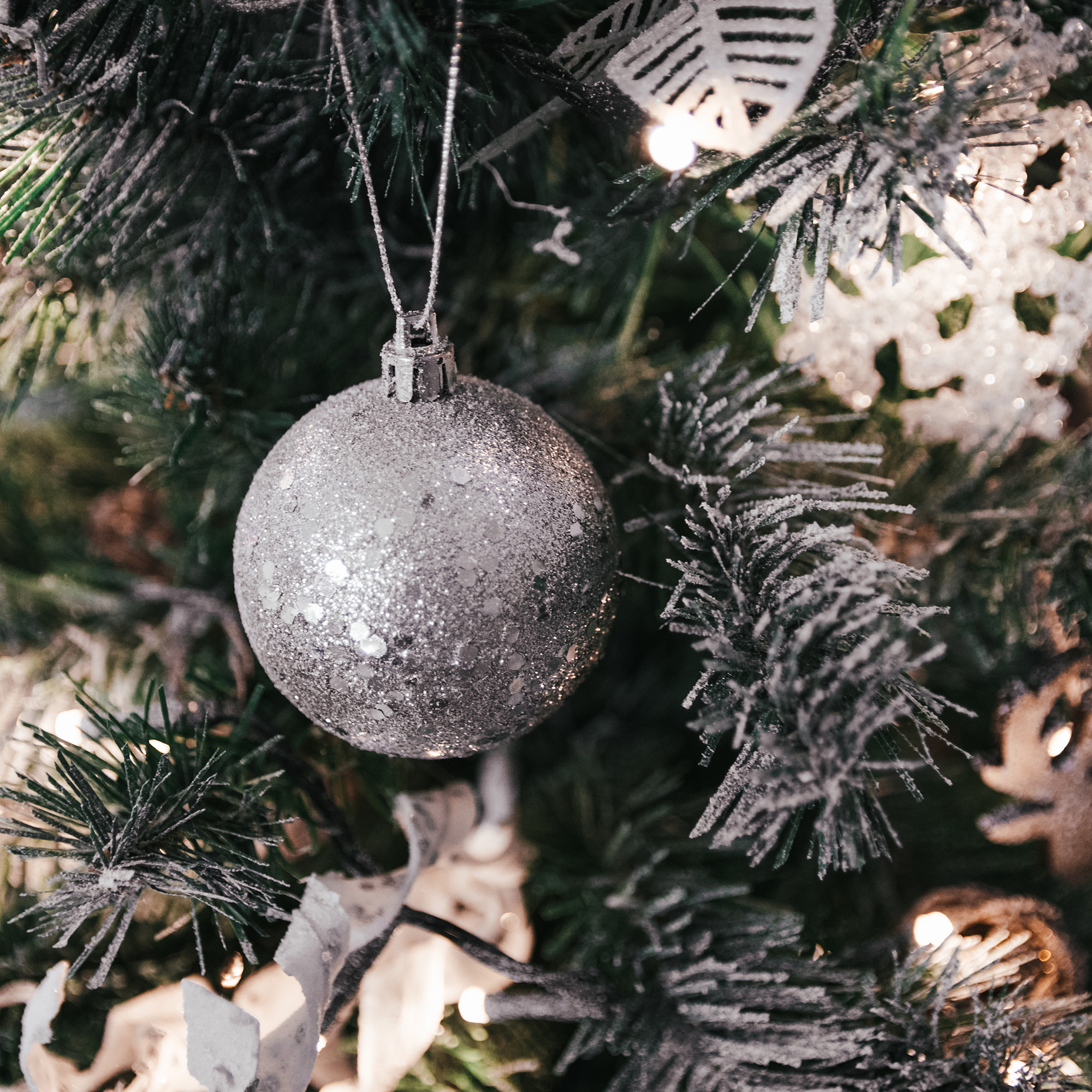 Primer plano de un árbol de Navidad. Una bola plateada con purpurina cuelga de una rama, con luces y adornos.