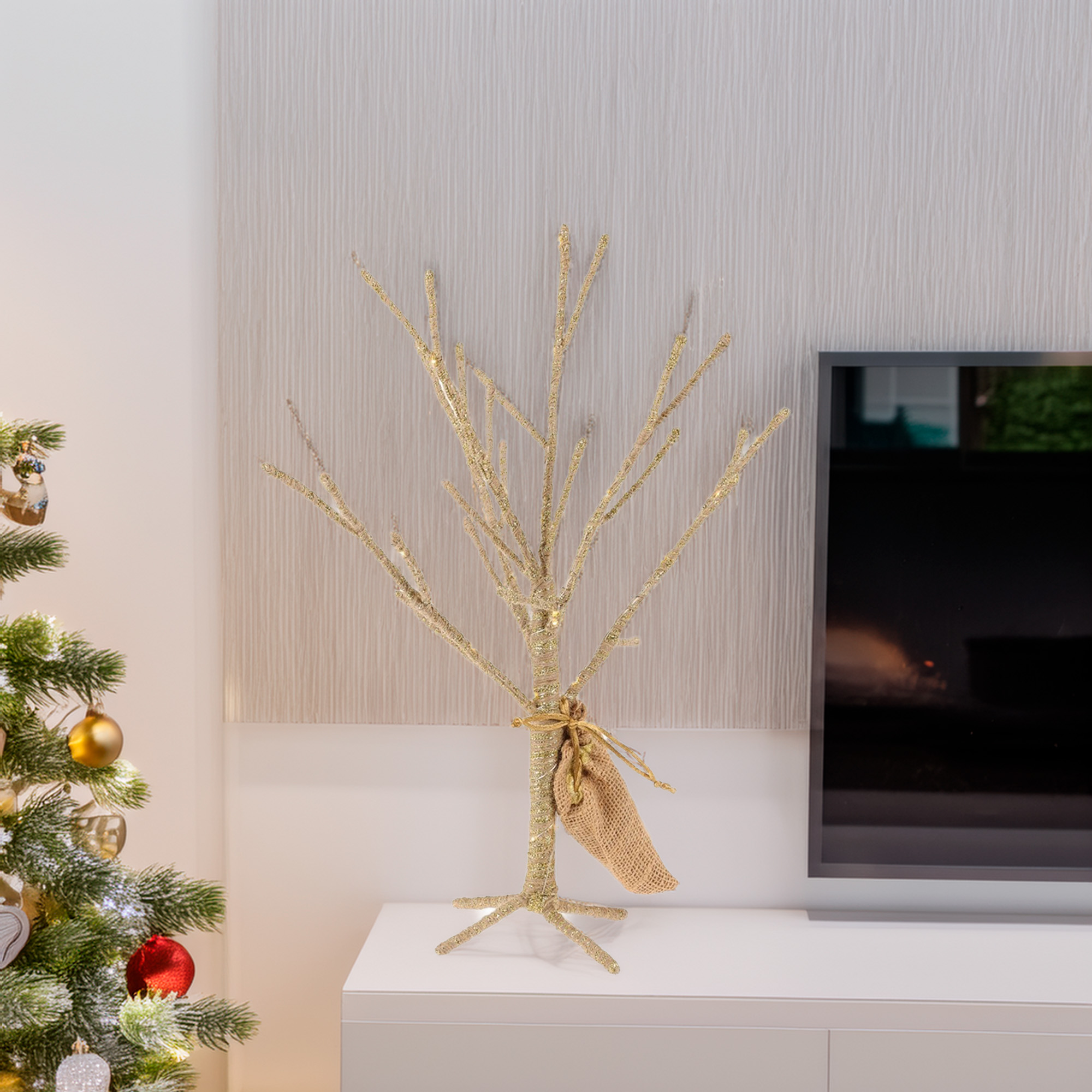 Un árbol decorativo dorado con un saco de arpillera está sobre una superficie blanca. Un árbol de Navidad a la izquierda.