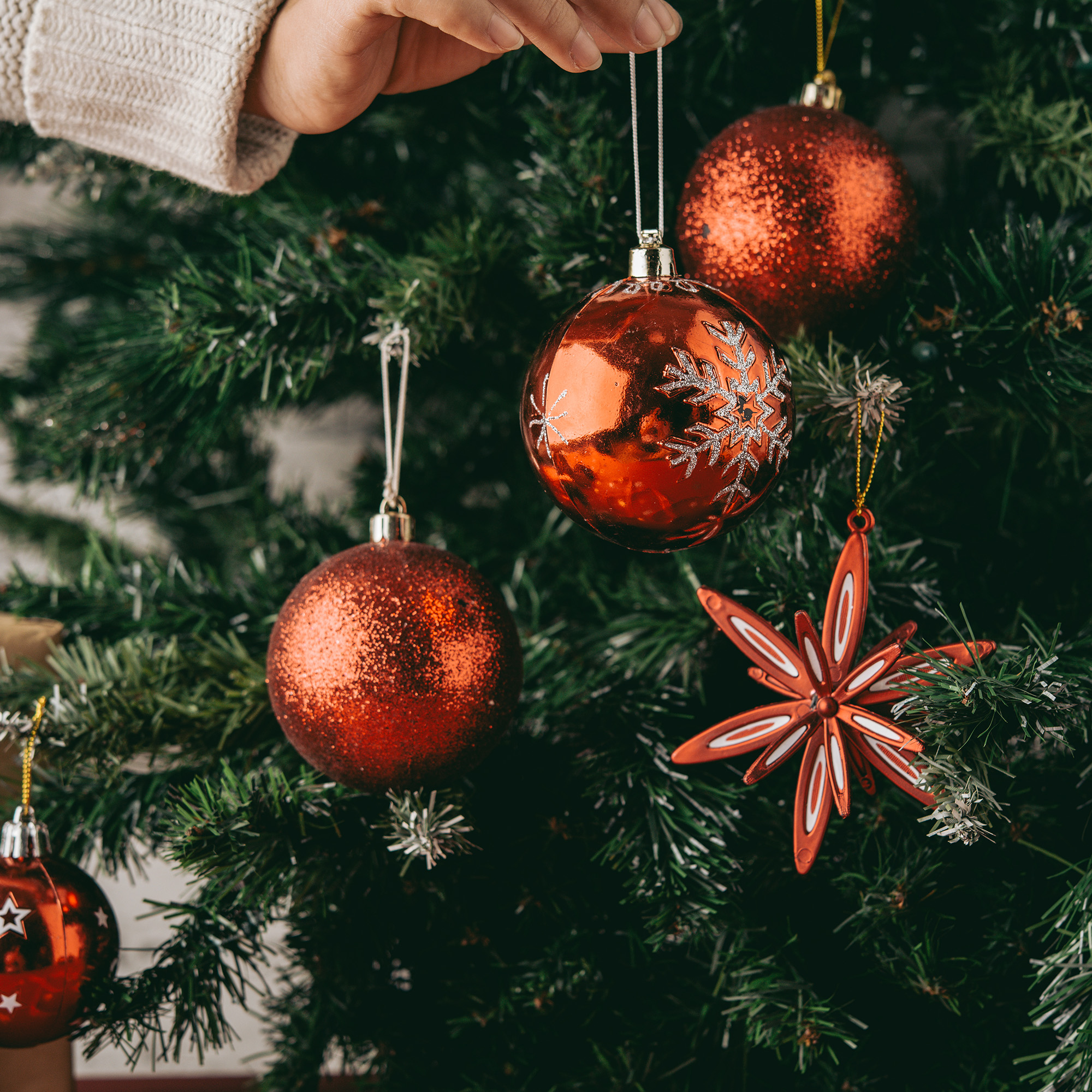 Una mano decora un árbol de Navidad con adornos rojos, incluyendo bolas y una decoración en forma de estrella.