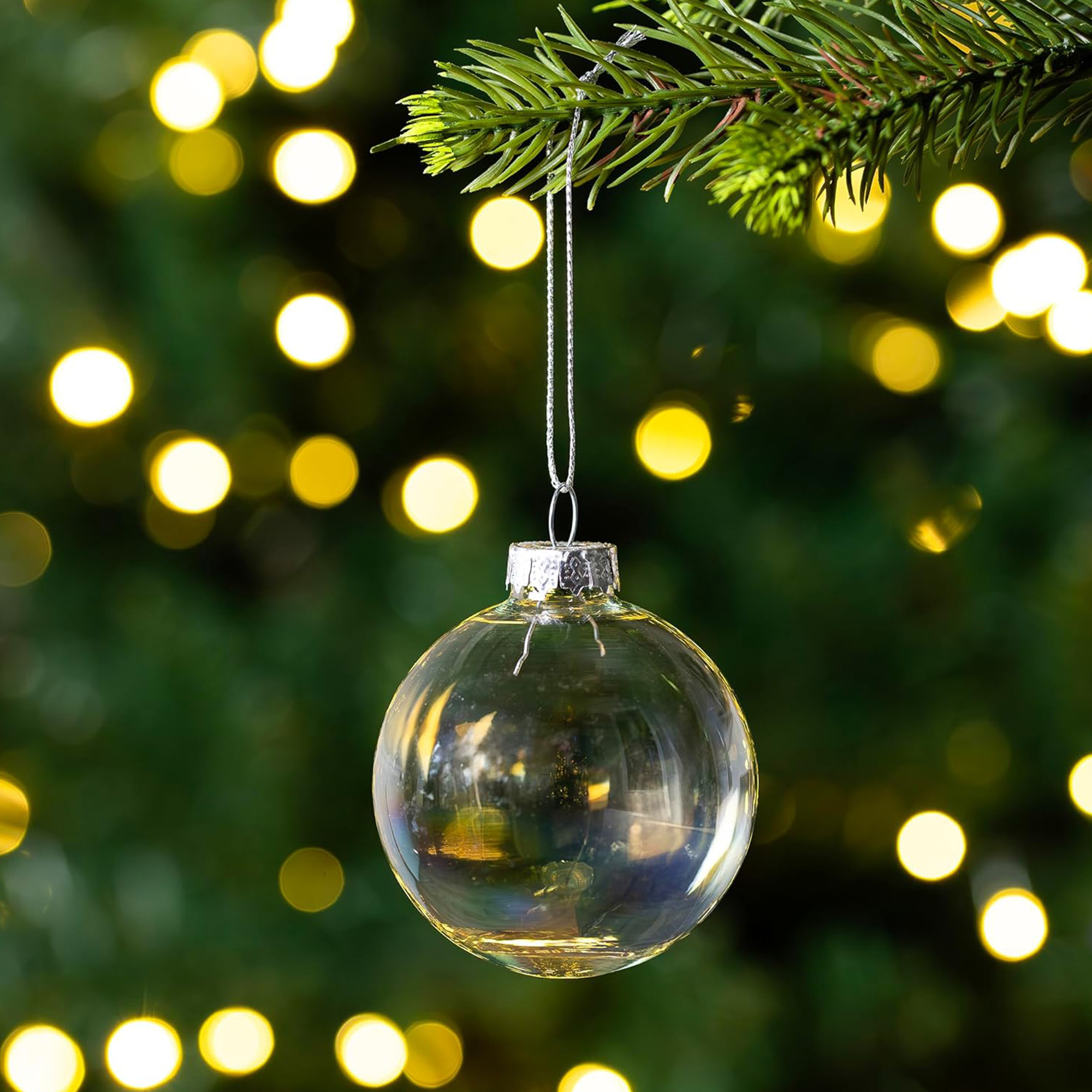 Un adorno navideño transparente cuelga de una rama. El fondo tiene luces borrosas.