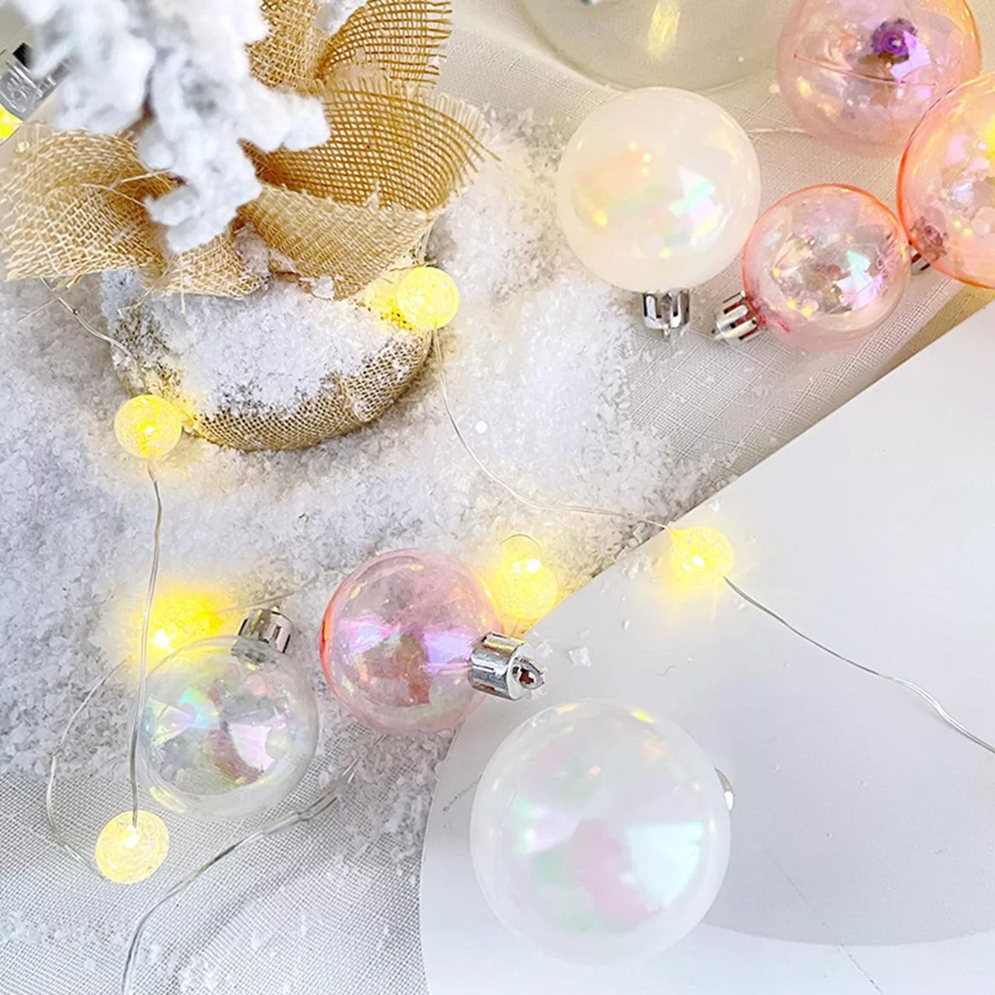 Adornos navideños sobre una superficie blanca con nieve artificial. Algunos son rosas y transparentes, otros blancos. Luces de hadas visibles.