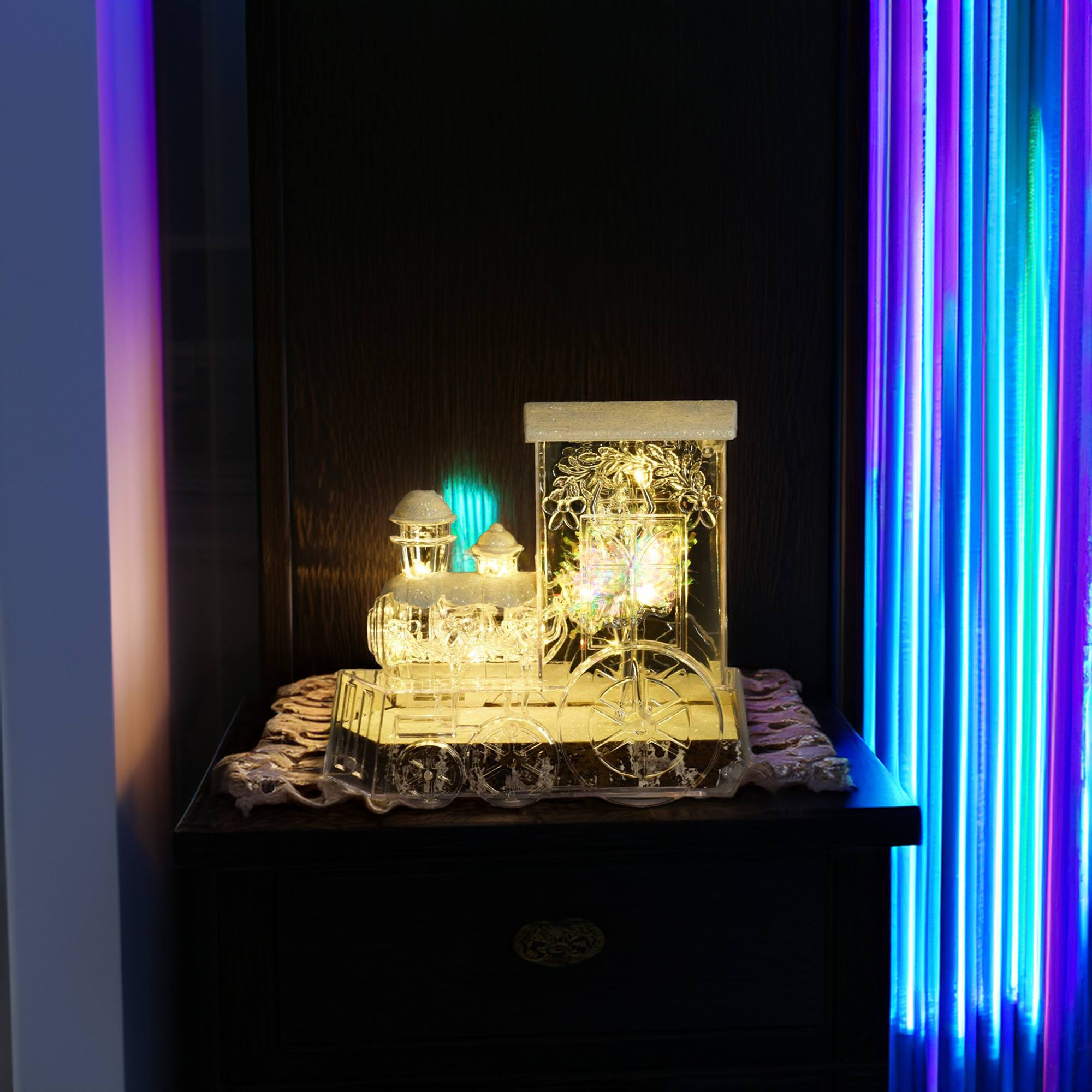 Una decoración de tren de plástico transparente iluminada descansa sobre una superficie oscura. Luces de neón coloridas brillan a la derecha.