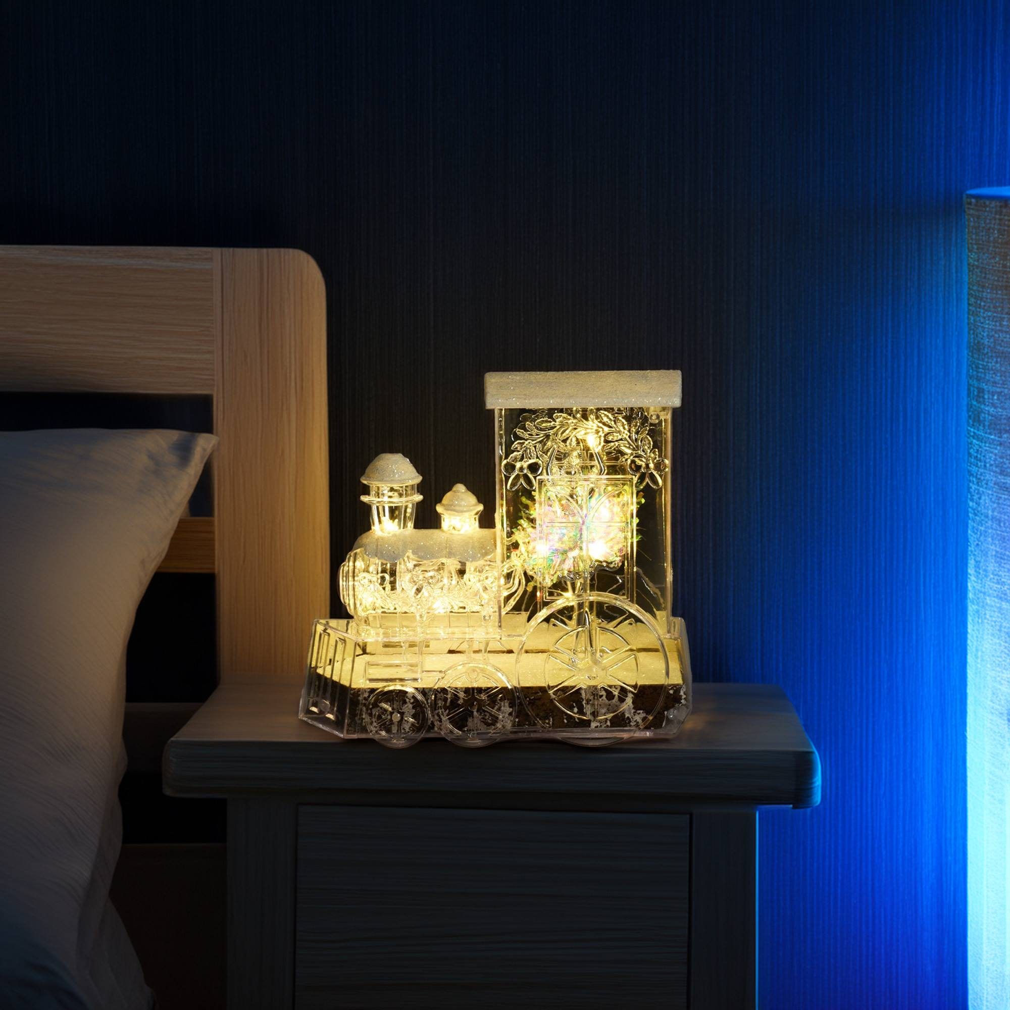 Una decoración en forma de tren iluminado está sobre una mesita de noche. Una cama a la izquierda, una lámpara a la derecha. Fondo oscuro.