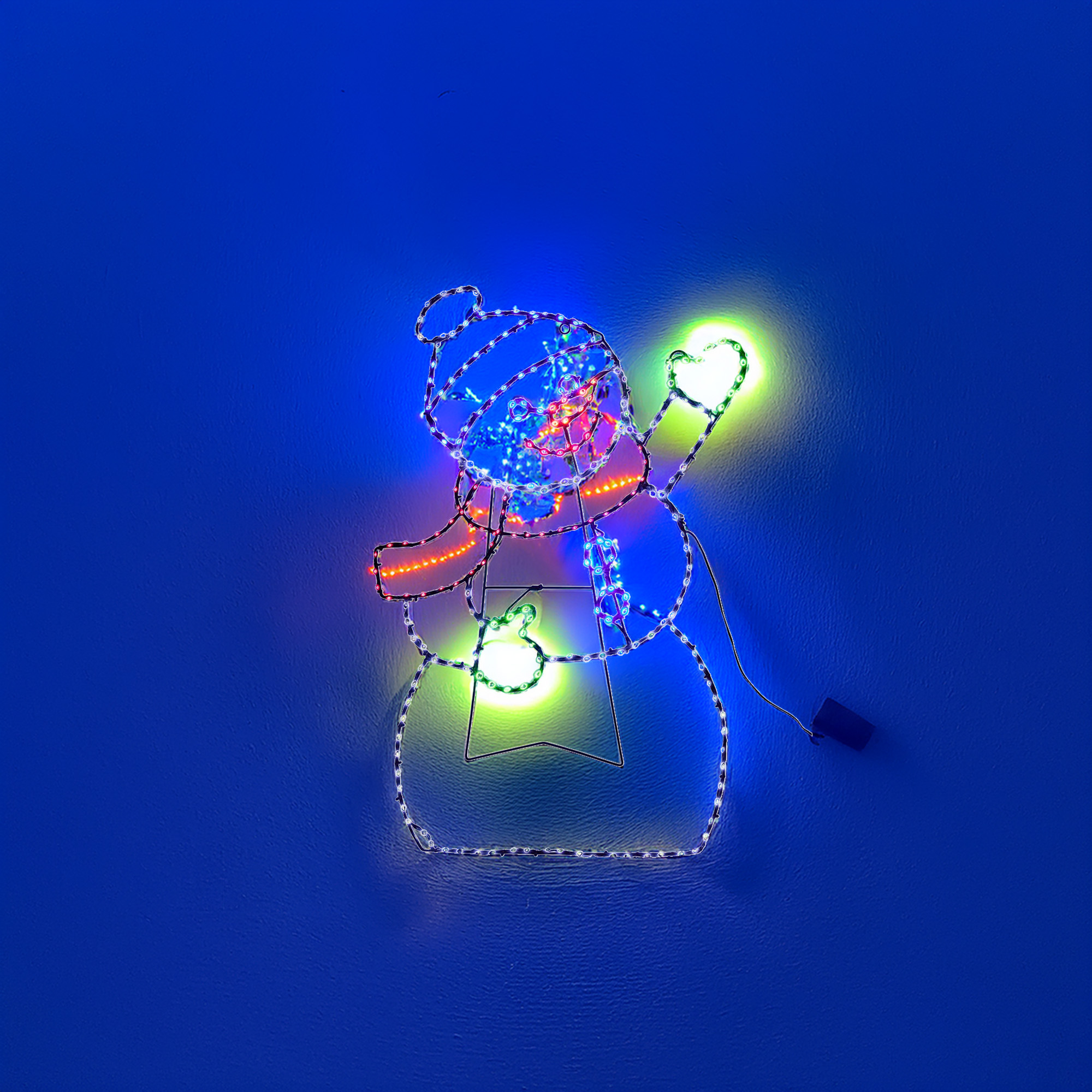 Una decoración navideña con forma de muñeco de nieve sobre una pared azul. Tiene sombrero, bufanda y brazos, todos iluminados.