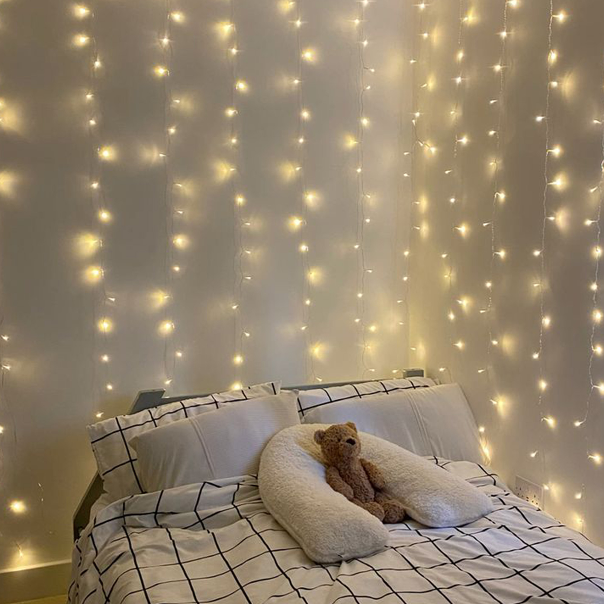 Un dormitorio con luces de hadas en la pared. Una cama tiene ropa de cama blanca y un pequeño oso de peluche.