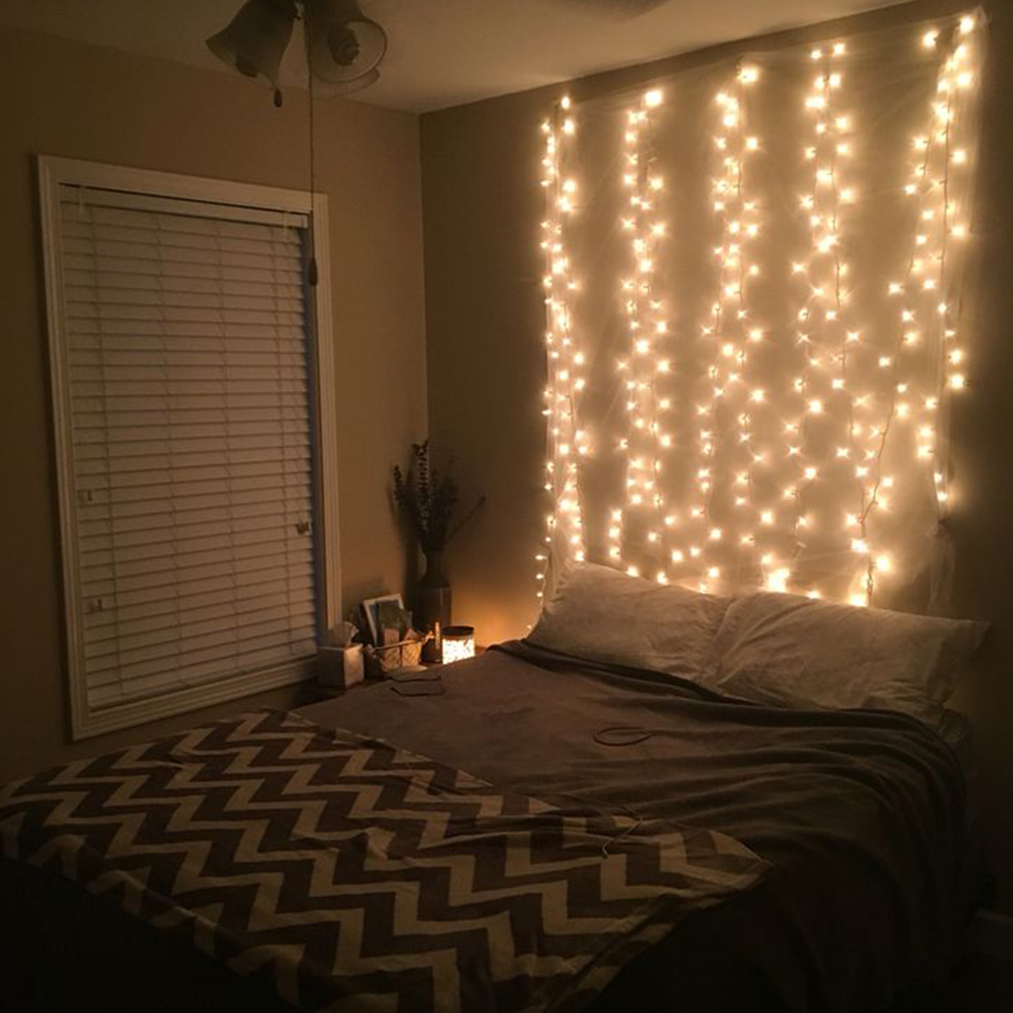 Un dormitorio con una cama, luces de hadas y una ventana con persianas. La habitación está tenue.