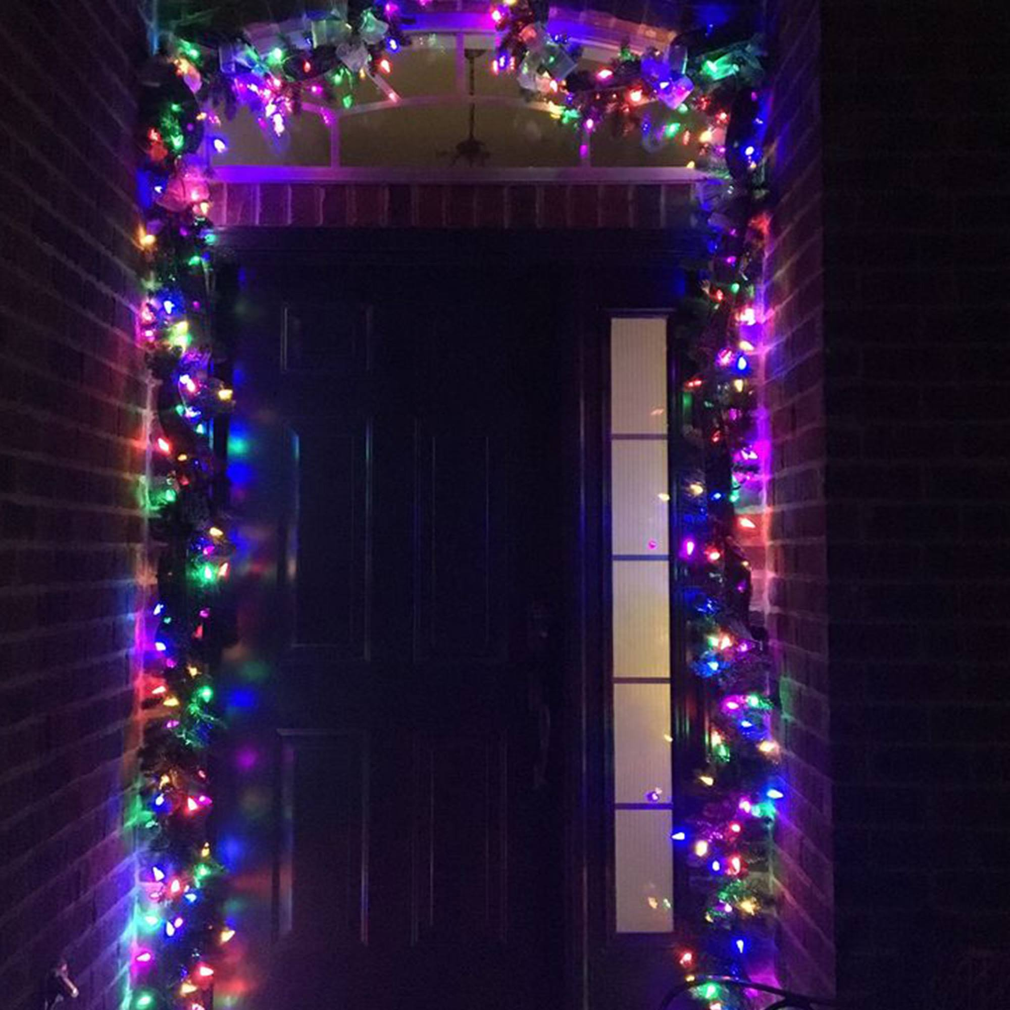 Una puerta principal oscura con luces navideñas y guirnalda. Pared de ladrillo a los lados. Las luces son multicolores.