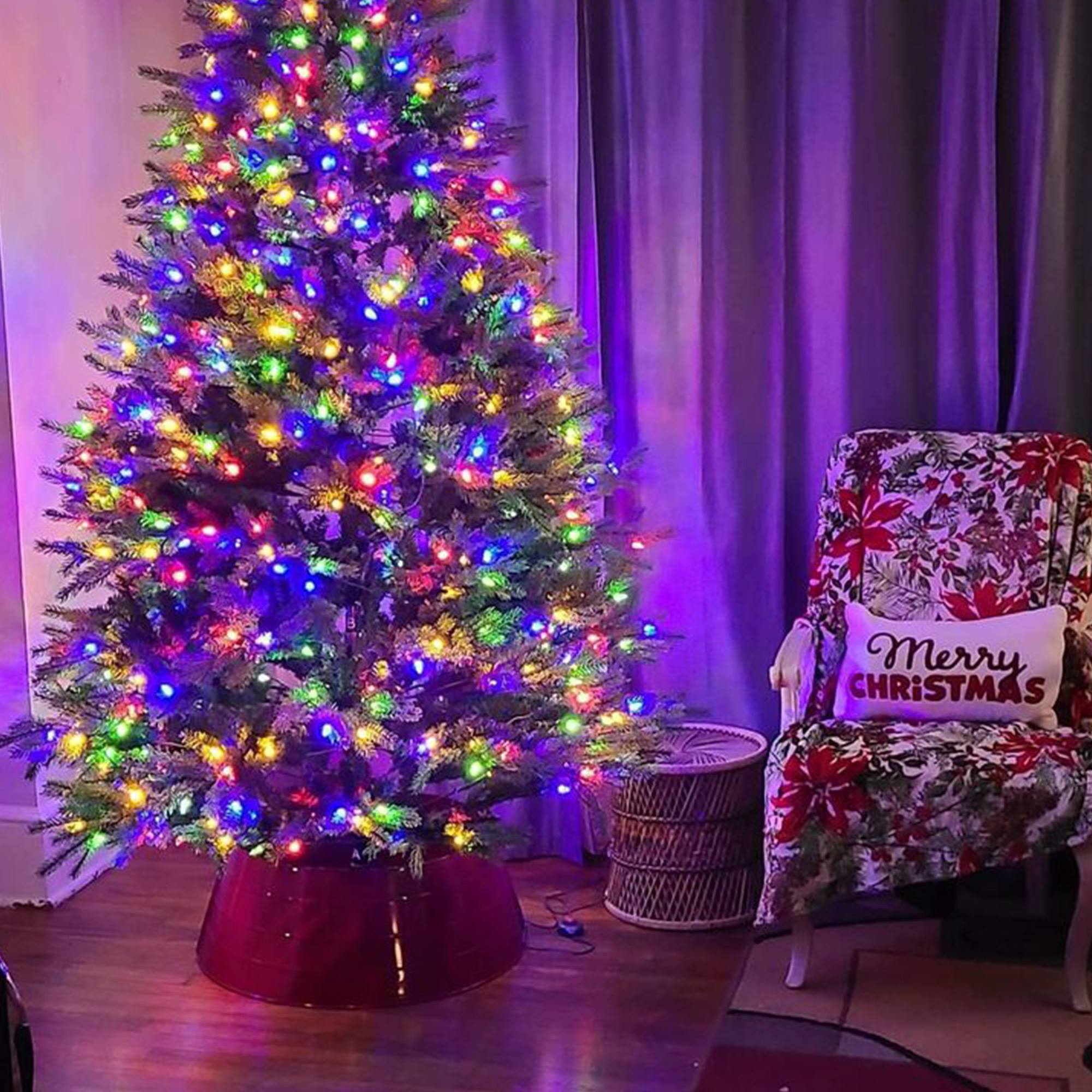 Un árbol de Navidad decorado con luces de colores. Cerca hay una silla con una almohada que dice 'Feliz Navidad'.