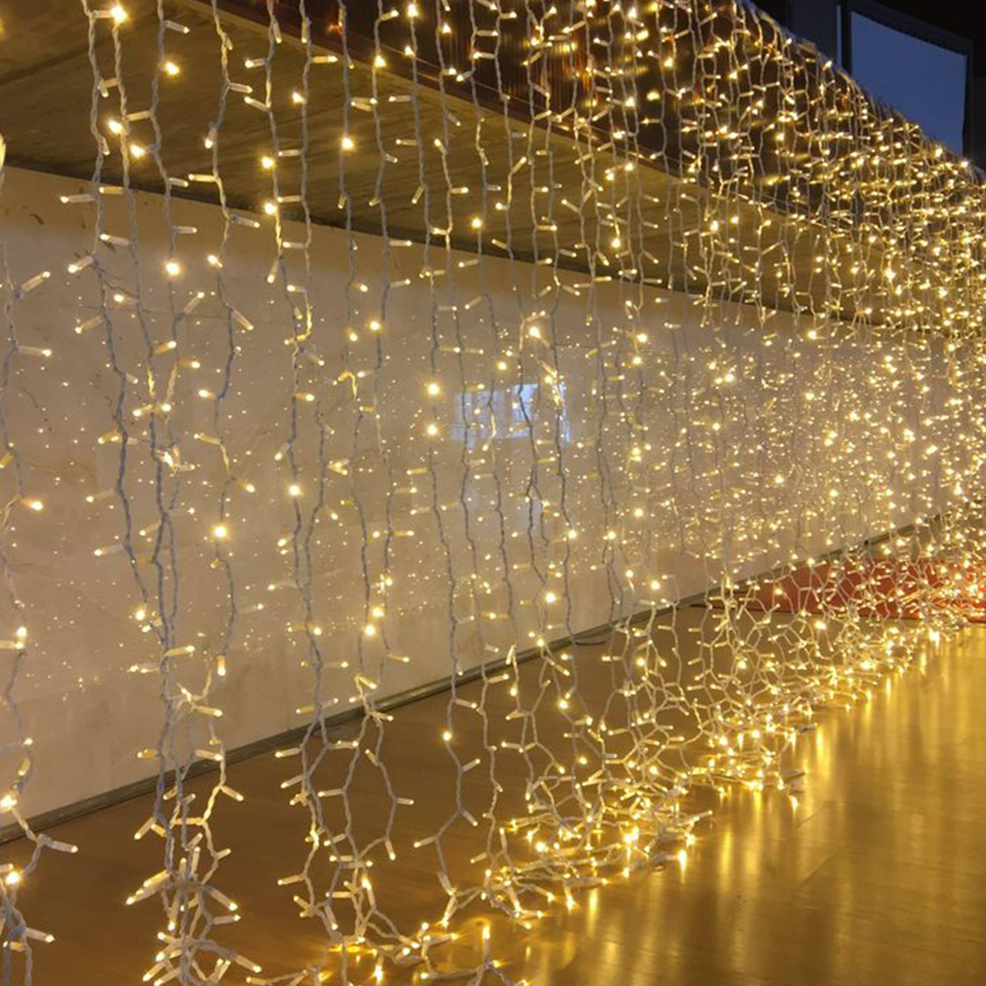 Una pared de luces blancas brillantes y cálidas. Las luces cuelgan desde arriba, creando un efecto de cortina. El suelo de madera refleja la luz.