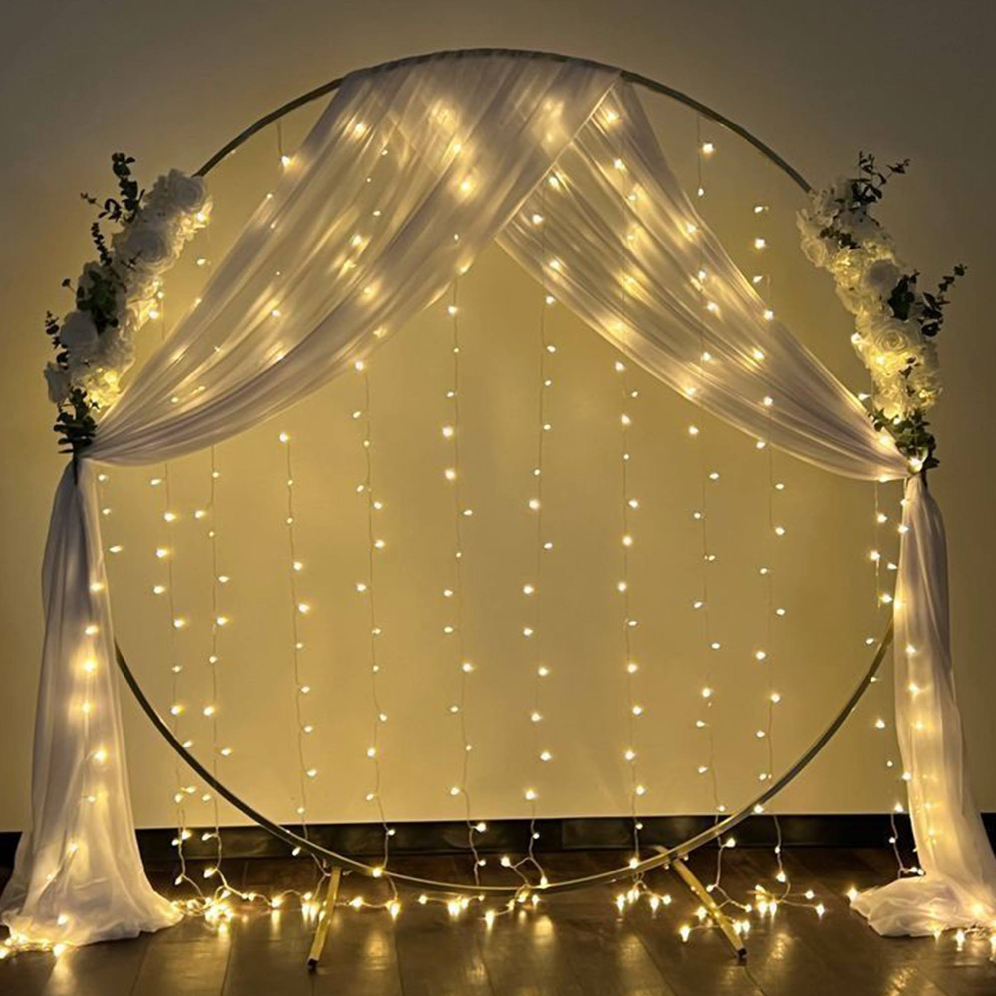 Un arco circular dorado decorado con tela blanca, luces de hadas y arreglos florales, creando un ambiente romántico.