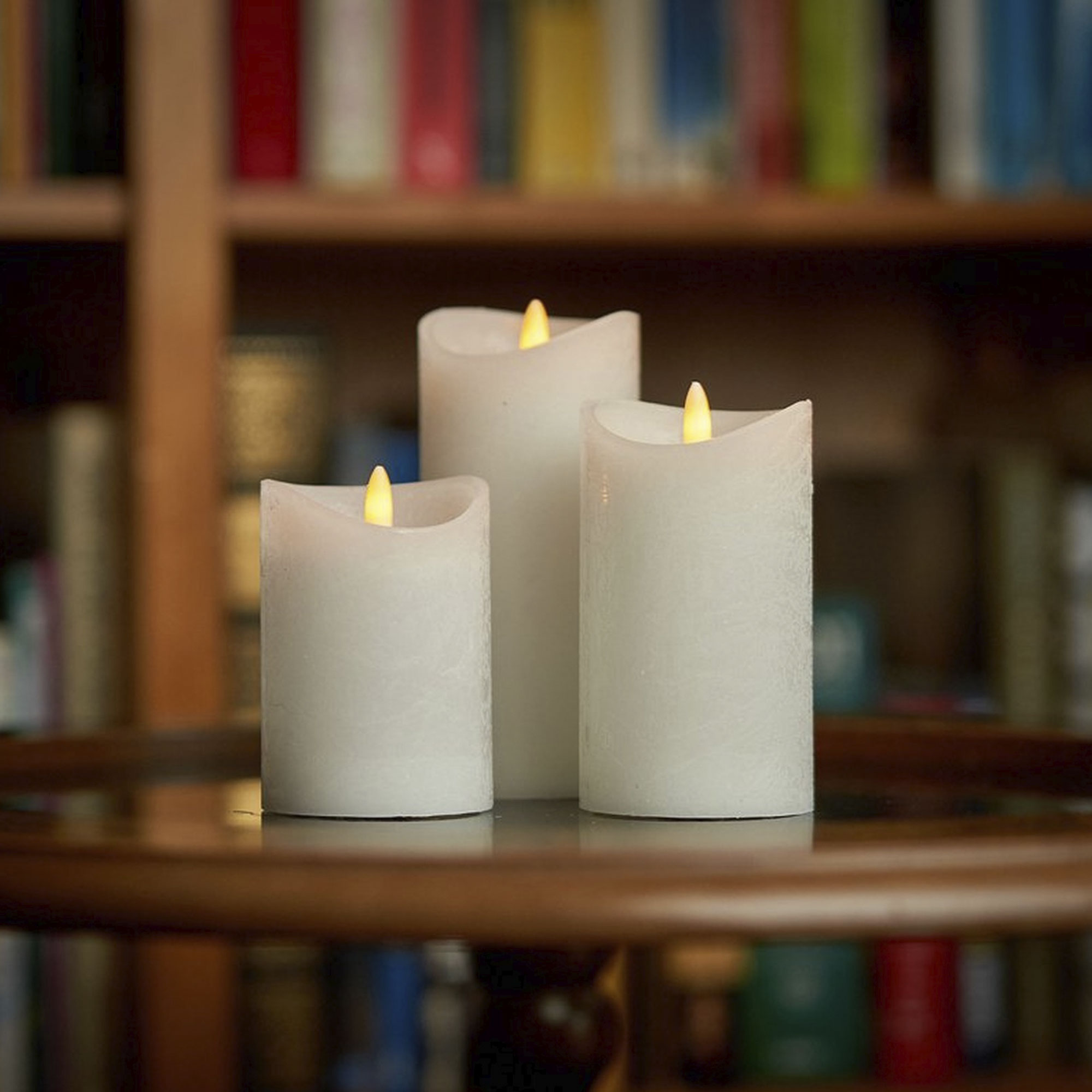 Tres velas blancas con llamas amarillas parpadeantes sobre una superficie reflectante, fondo de estantería.