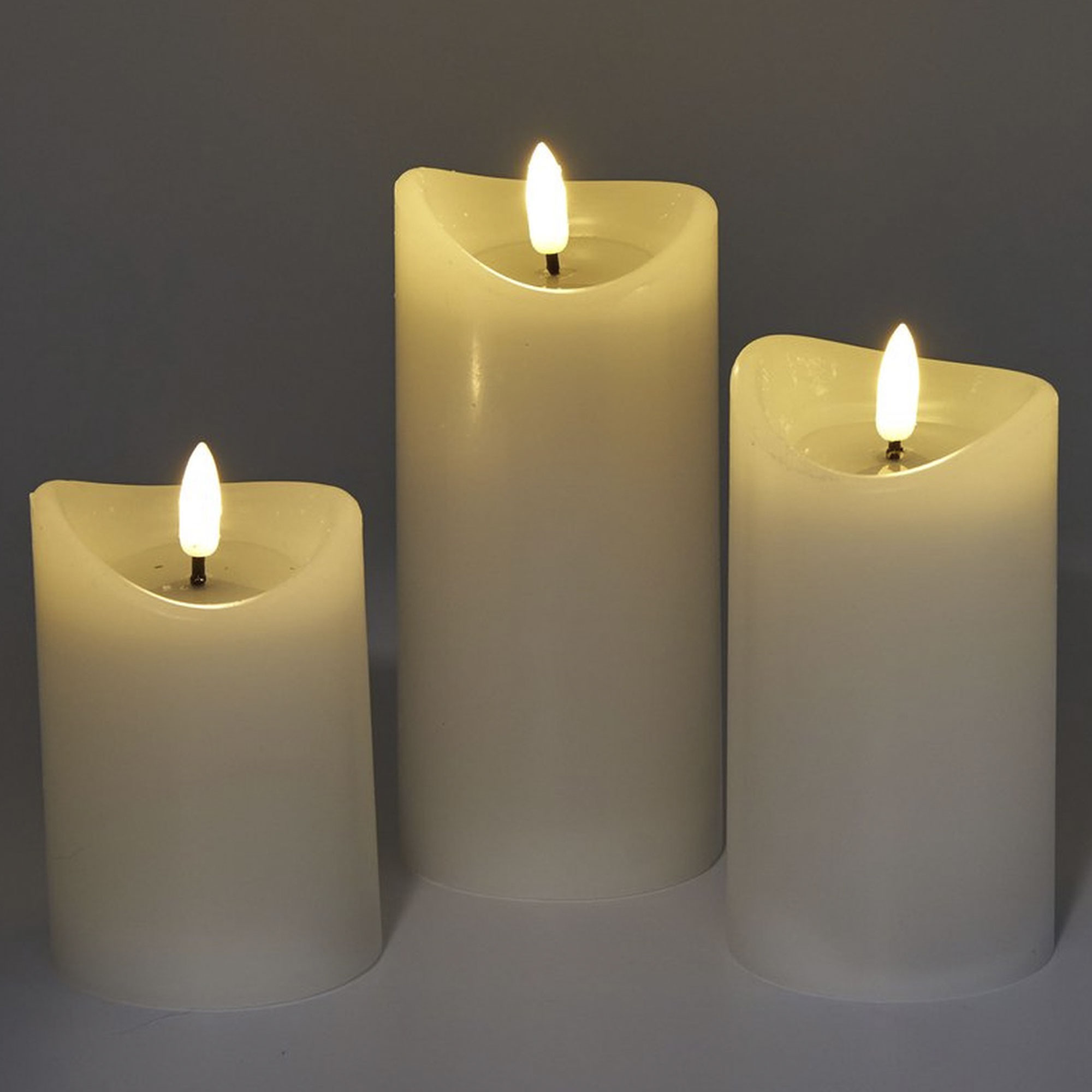 Tres velas marfil de diferentes alturas, cada una con una llama amarilla parpadeante, sobre un fondo gris.