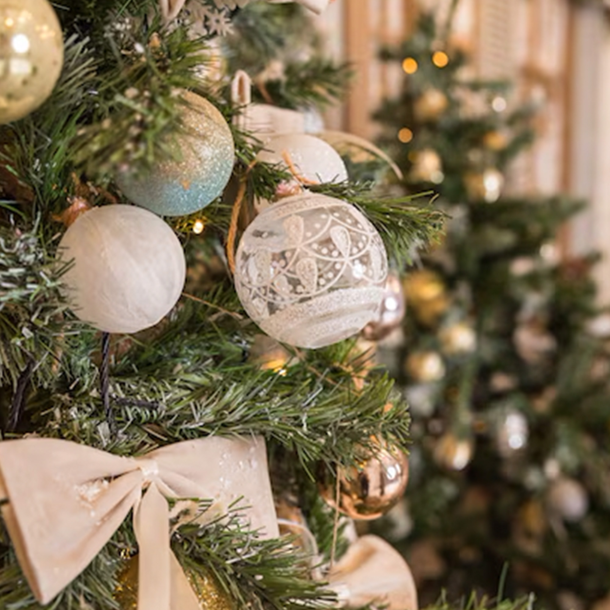 Primer plano de un árbol de Navidad decorado. Los adornos son blancos, dorados y azules, con un lazo crema. Otro árbol está al fondo.