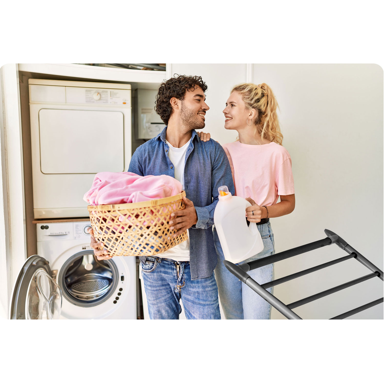 Para w pralni. Mężczyzna trzyma kosz z praniem, kobieta butelkę z detergentem. Widoczne pralka i suszarka. Patrzą na siebie.