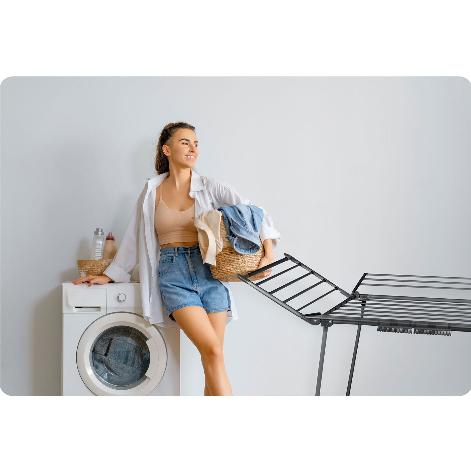 Kobieta opiera się o pralkę, trzymając kosz z praniem. Suszarka na pranie po prawej. Butelki i kosz na pralce.