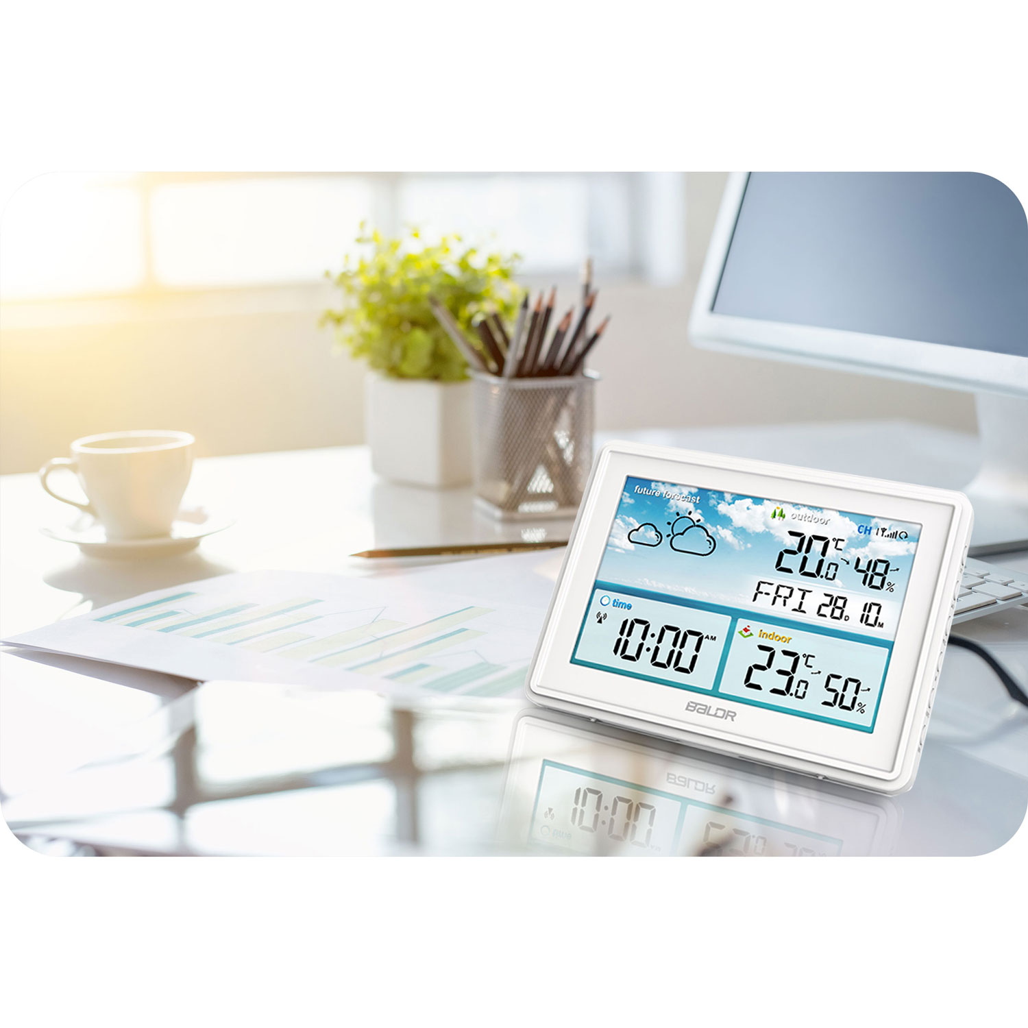 Biała cyfrowa stacja pogodowa wyświetla czas, temperaturę i prognozę pogody na biurku z filiżanką, roślinami i komputerem.