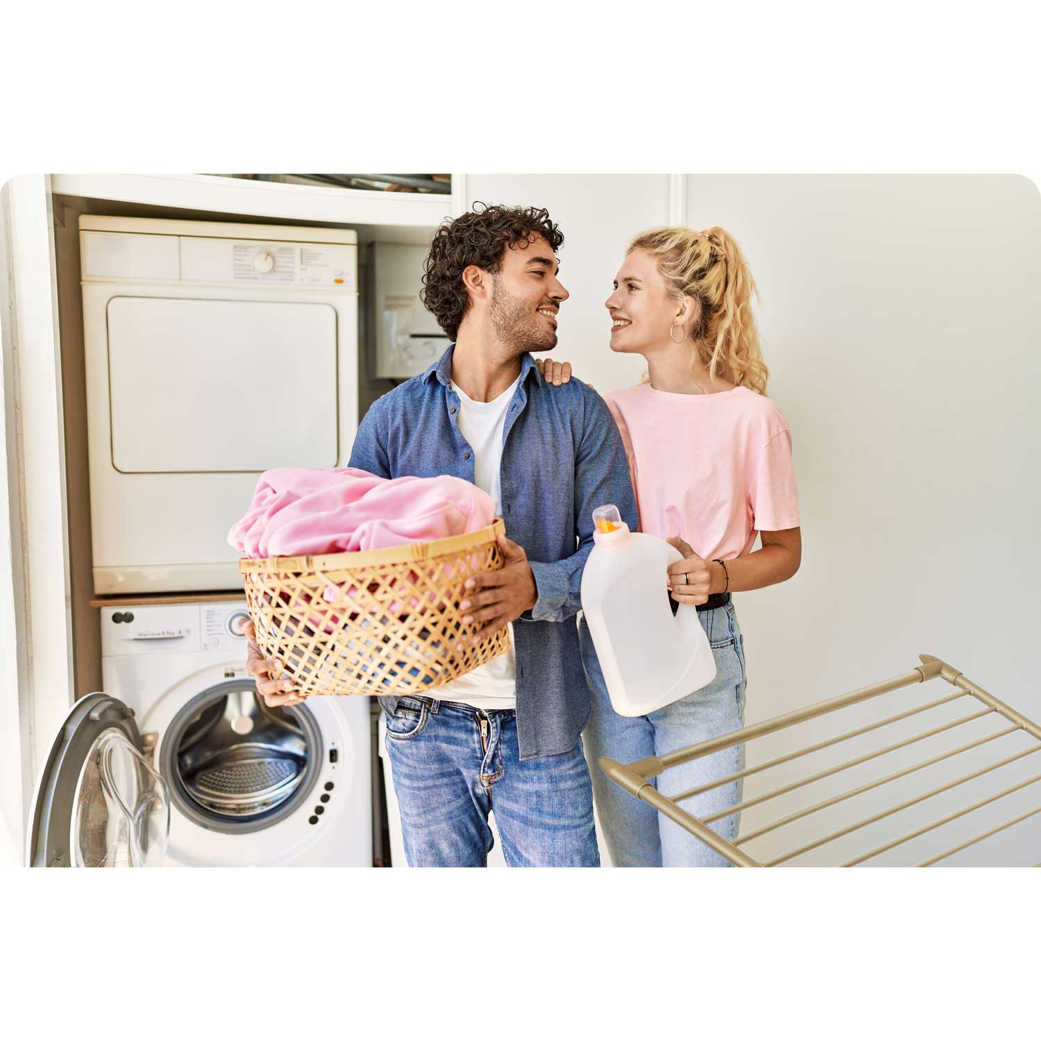 Para uśmiecha się do siebie w pralni. Mężczyzna trzyma kosz różowych ubrań, kobieta detergent, widoczne są pralki.
