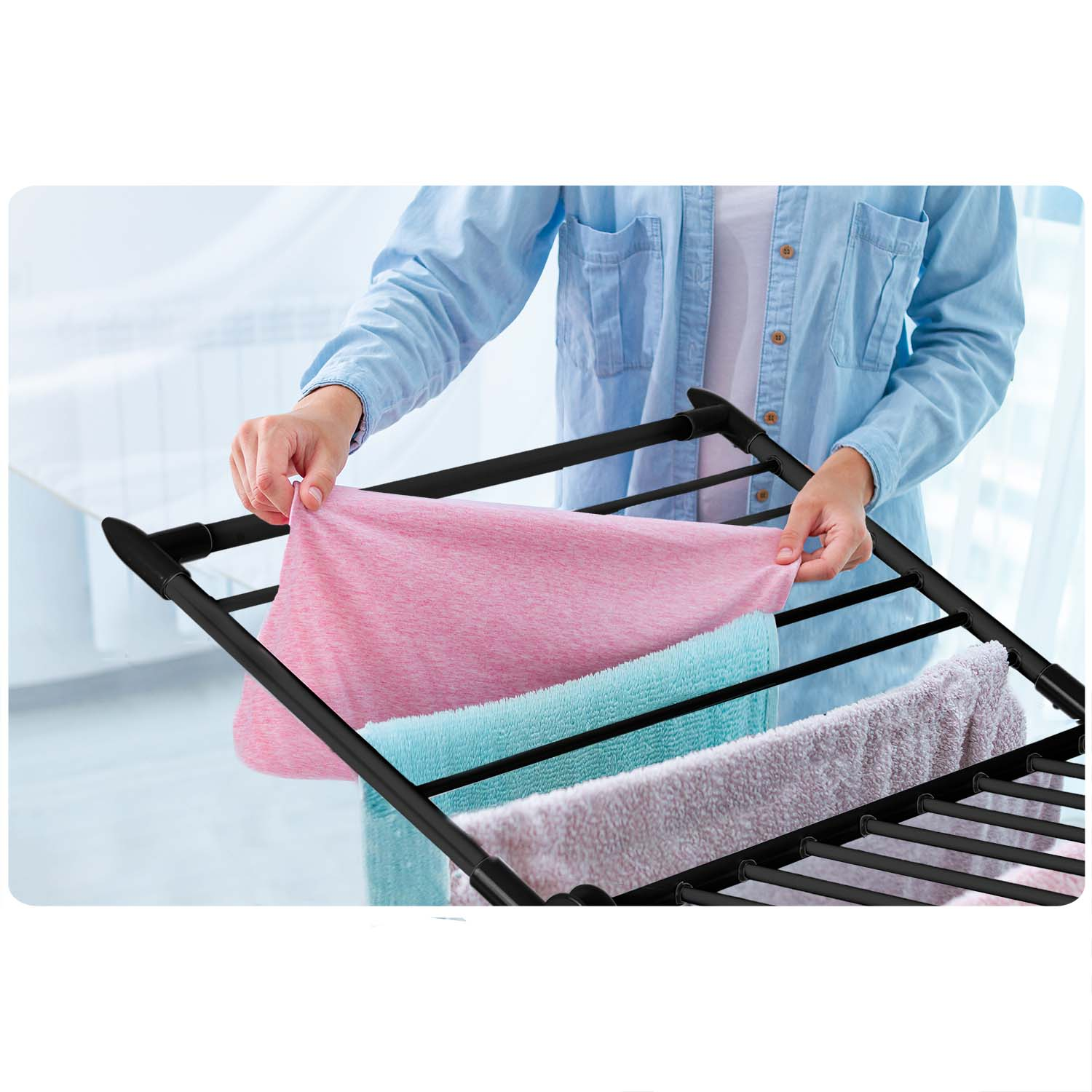 Osoba wiesza różowe, niebieskie i jasnofioletowe ręczniki na czarnym stojaku do suszenia. Osoba ma na sobie jasnoniebieską koszulę.