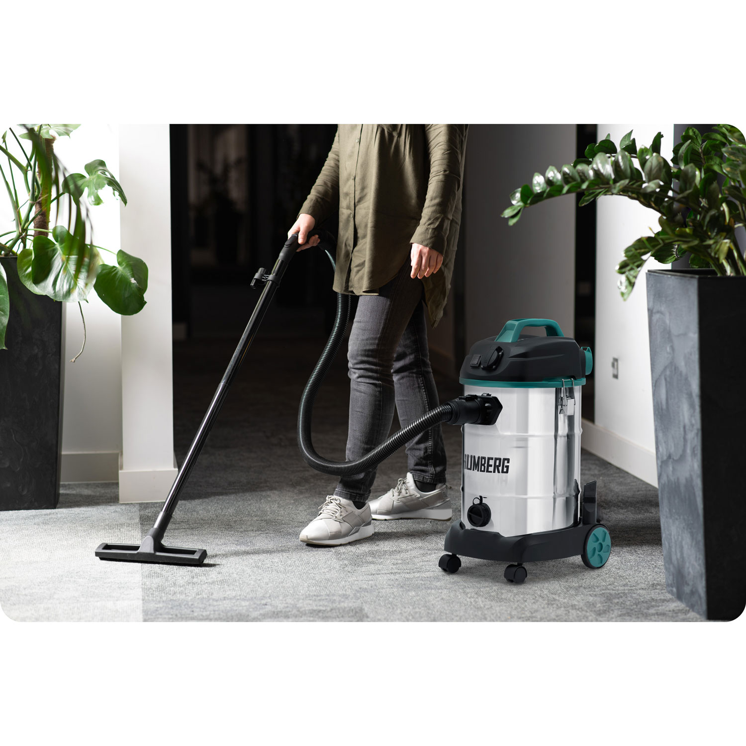 Osoba odkurza dywan na podłodze biurowej srebrnym odkurzaczem. Zielone rośliny doniczkowe znajdują się po obu stronach obszaru odkurzania.