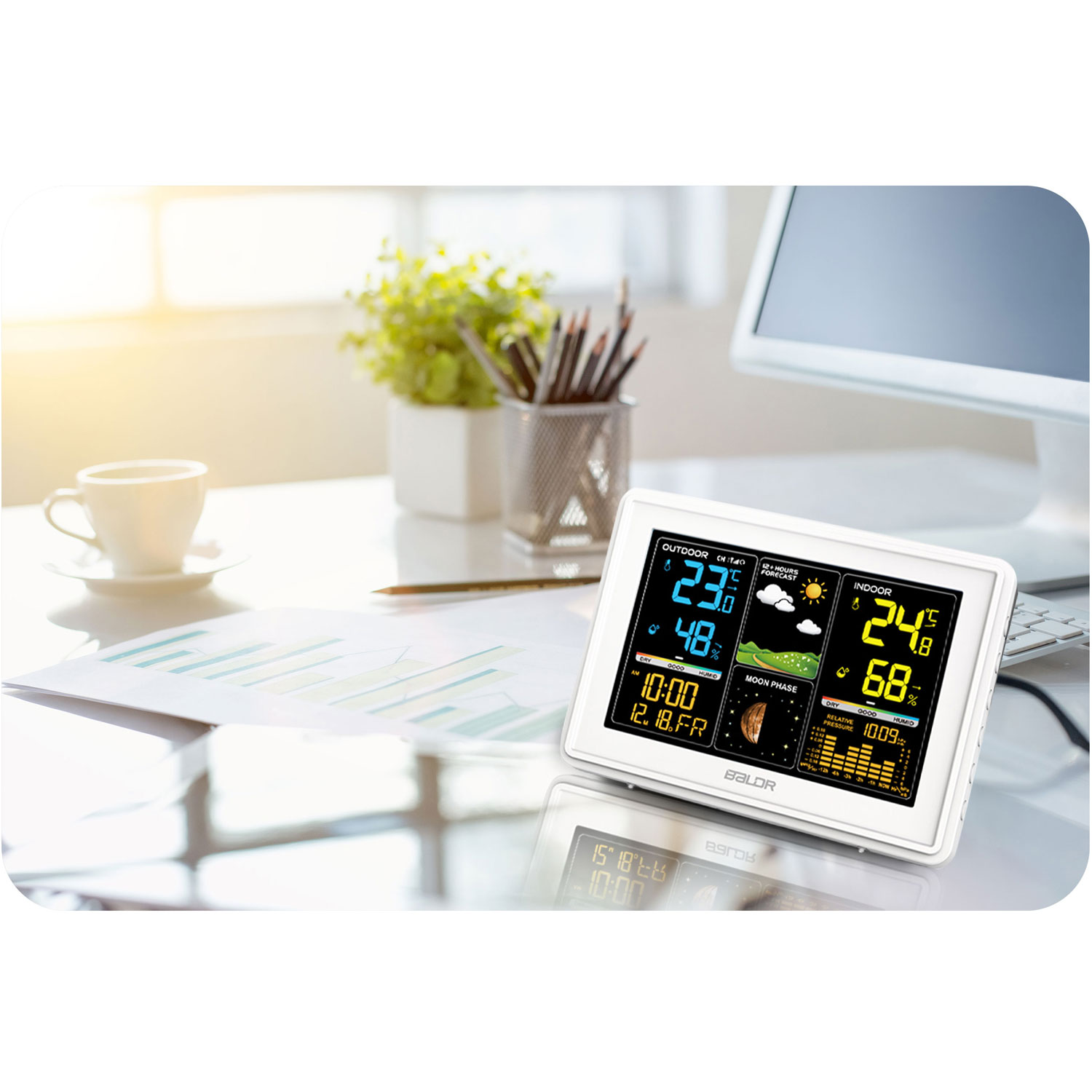 Stacja pogodowa na biurku wyświetla temperatury zewnętrzne/wewnętrzne, wilgotność, czas i prognozę pogody. Widoczny jest również kubek, rośliny i komputer.