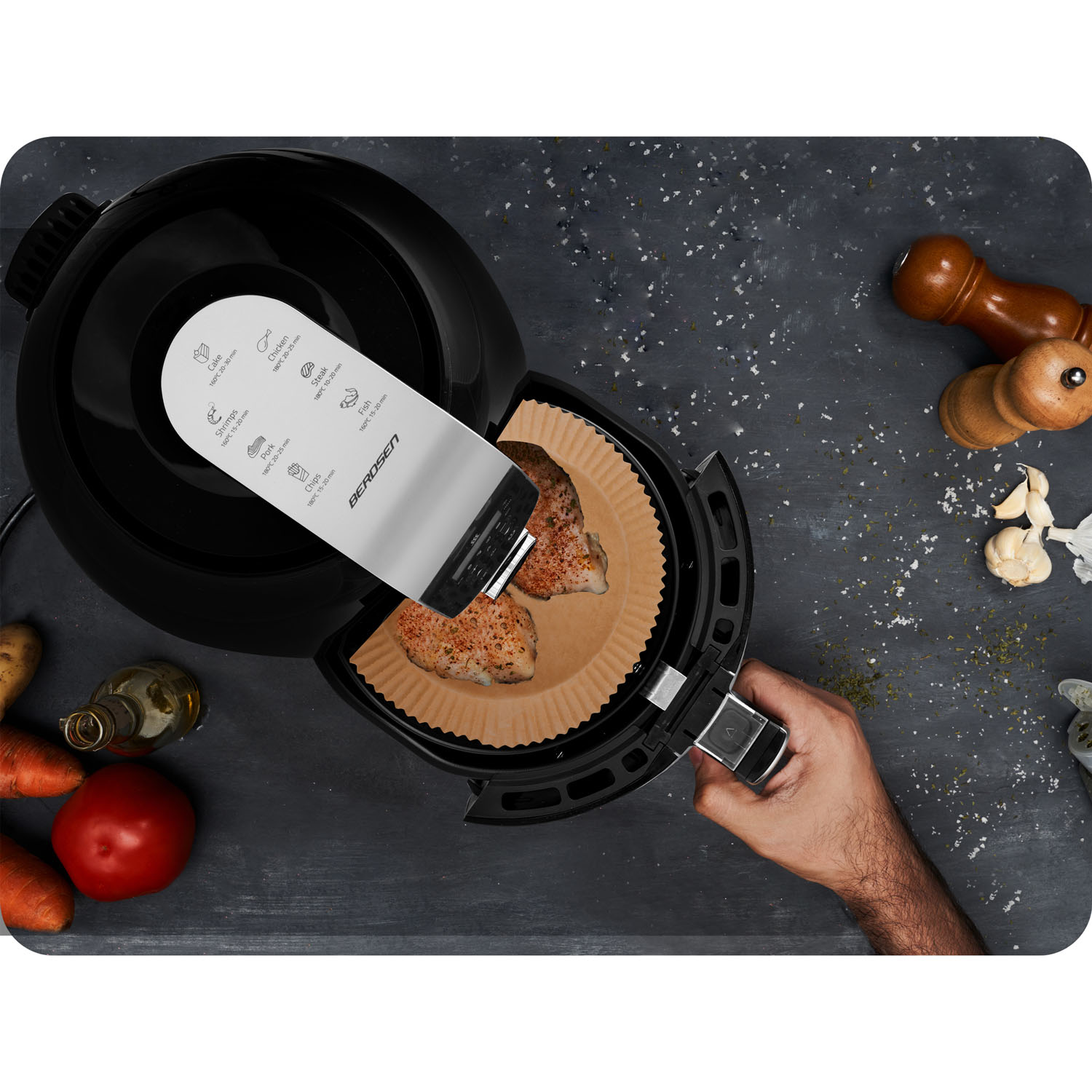 Otwarta frytkownica powietrzna z jedzeniem w środku, dłoń trzymająca ją i warzywa na ciemnej powierzchni.