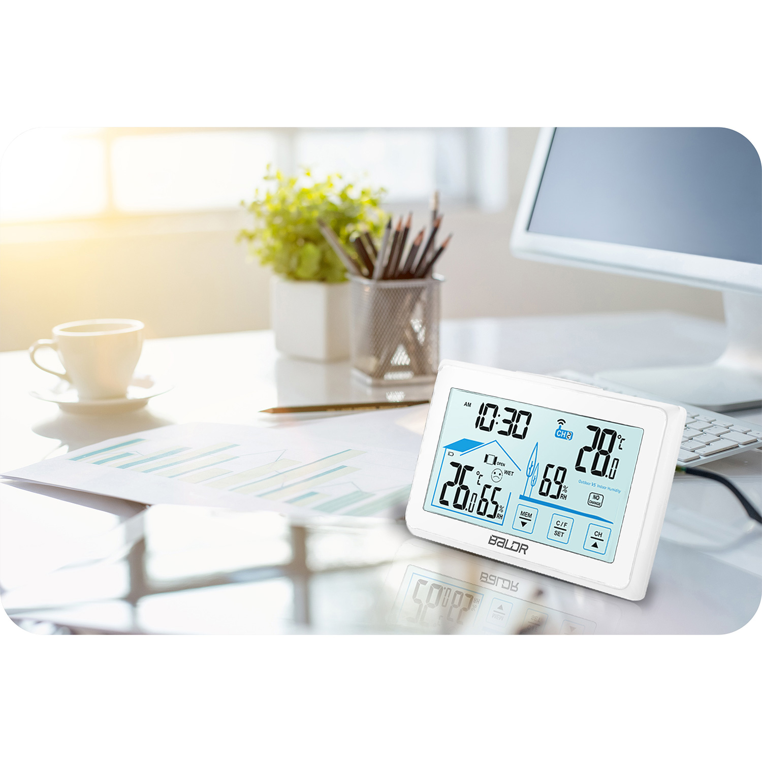 Białe biurko ze stacją pogodową pokazującą godzinę, temperaturę i wilgotność. W tle filiżanka kawy, rośliny i komputer.