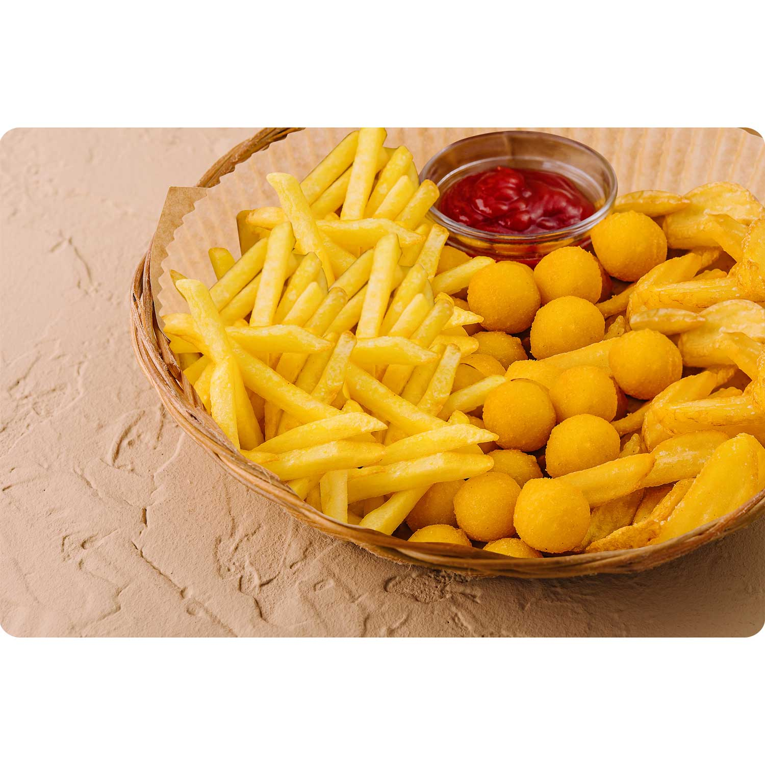 Koszyk zawiera frytki, kulki serowe i małą szklaną miseczkę z ketchupem. Jedzenie znajduje się na teksturowanej beżowej powierzchni.