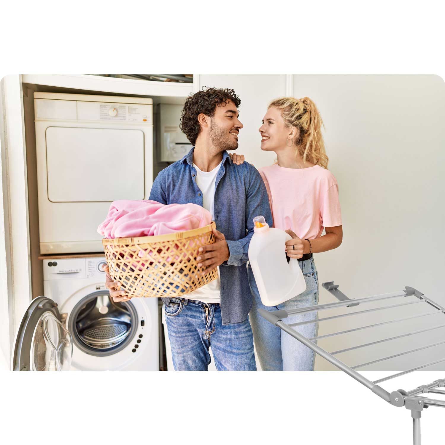 Para w pralni, mężczyzna trzymający kosz różowych ubrań, kobieta trzymająca detergent. Pralki i suszarka w tle.