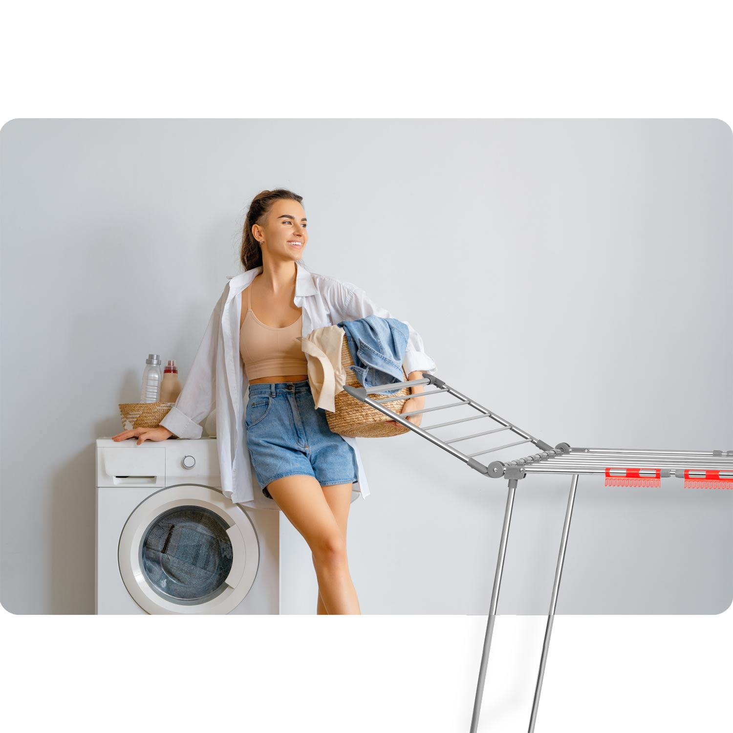 Kobieta z koszem na pranie opiera się o pralkę. Suszarka po prawej. Neutralne tło.