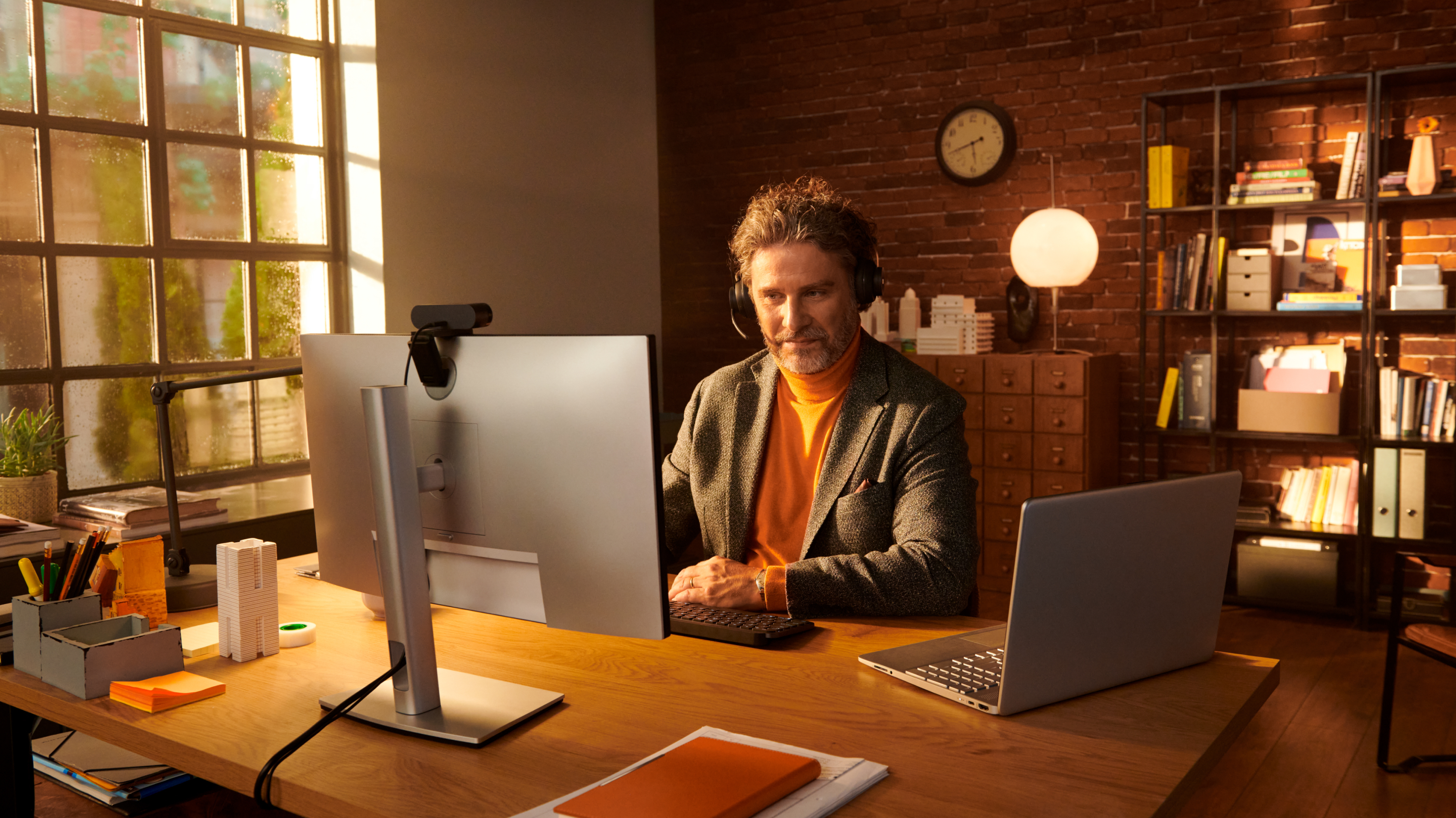 Un uomo in giacca e dolcevita arancione lavora a una scrivania con un monitor. Laptop, orologio e scaffali di libri sono in vista.