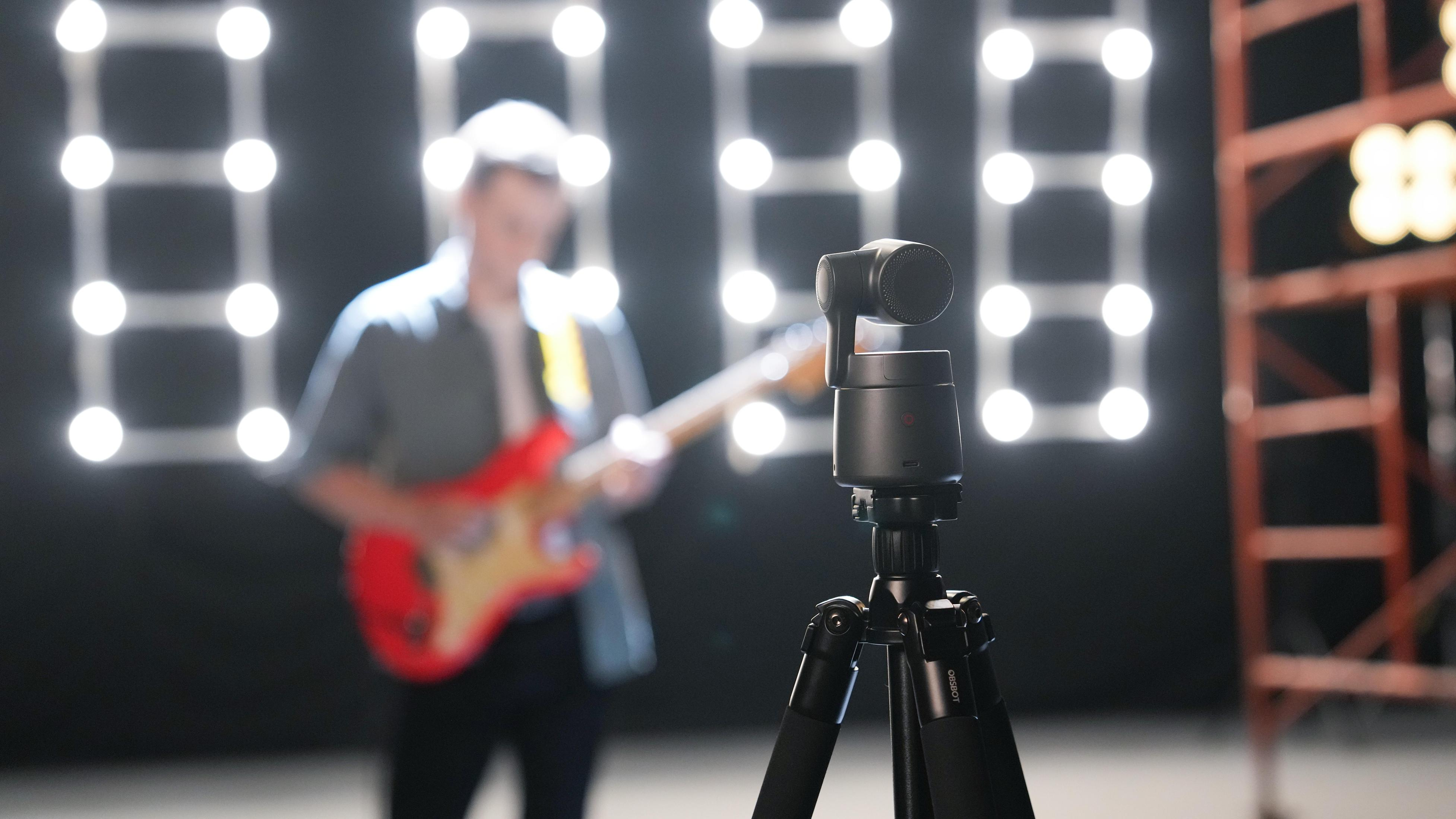 Kamera na statywie jest ustawiona do nagrywania muzyka grającego na czerwonej gitarze. W tle światła.