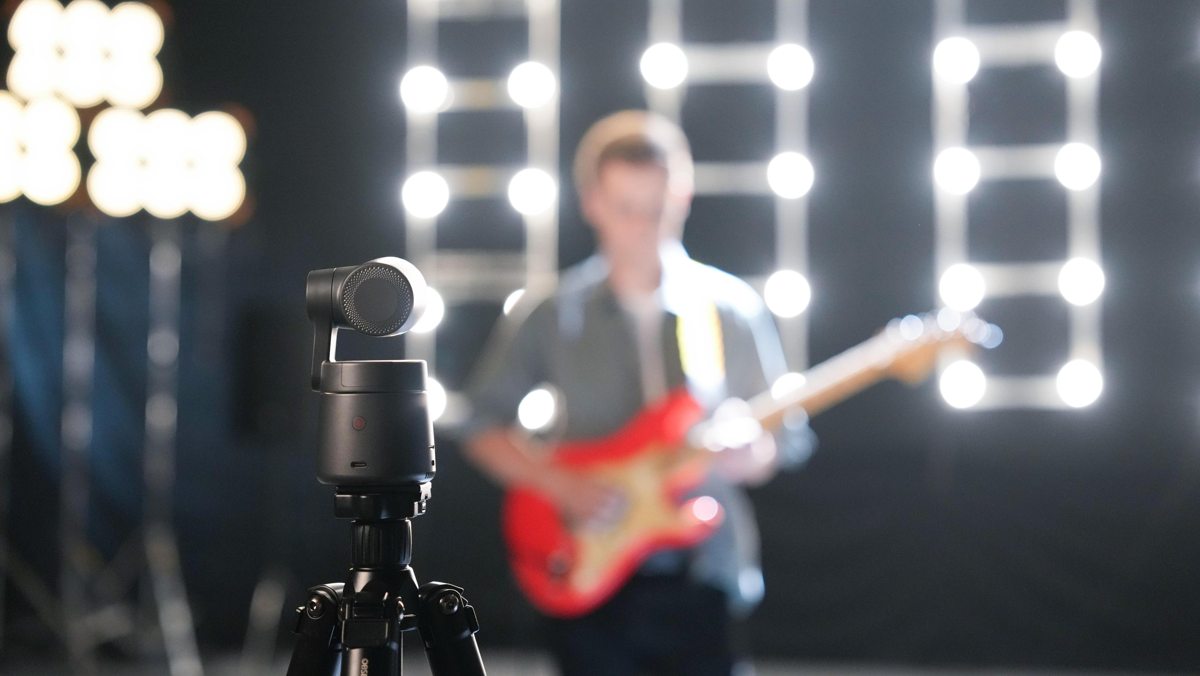 Kamera na statywie w ostrości. Osoba grająca na czerwonej gitarze jest rozmyta w tle, ze światłami.