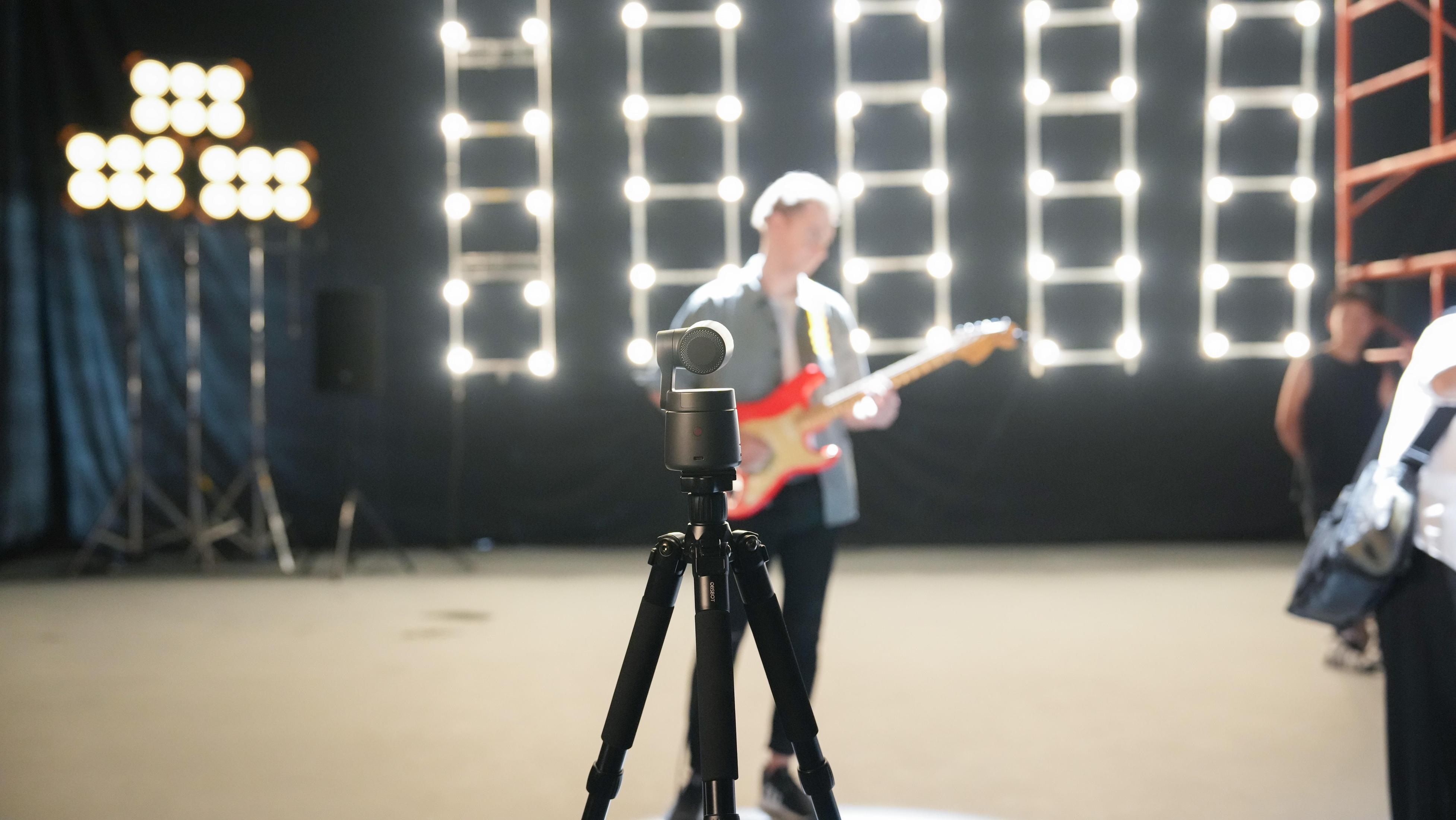 Kamera na statywie na pierwszym planie. Osoba grająca na gitarze w tle, światła z tyłu.