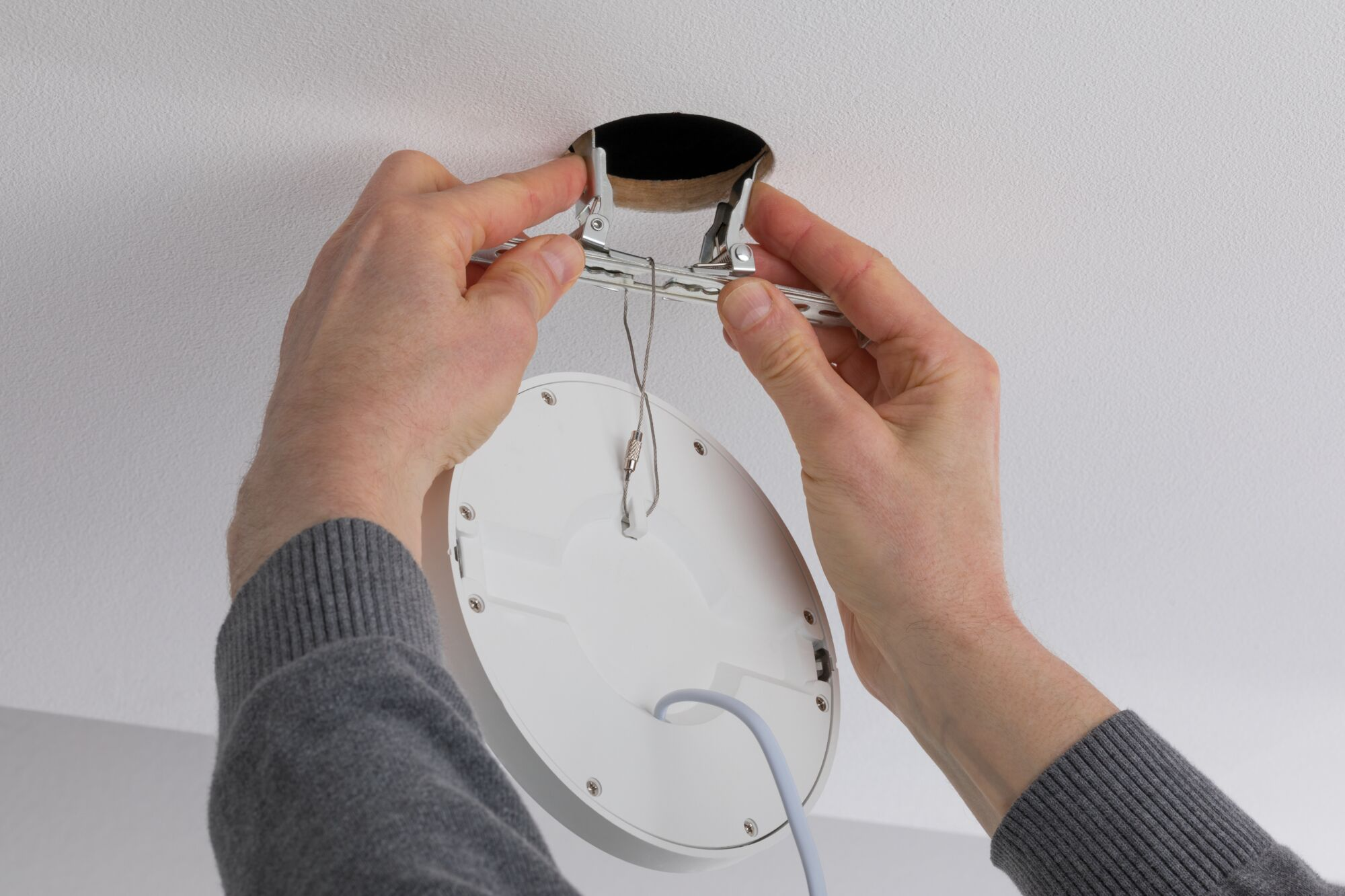 Eine Person installiert eine weiße Deckenleuchte. Hände halten die Leuchte und die Halterungen und führen sie in ein Deckenloch ein. Ein graues Kabel hängt.