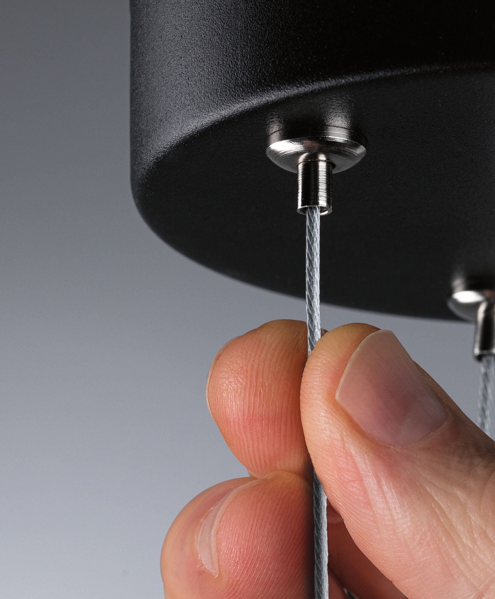 Dłoń trzyma stalową linkę połączoną z czarną oprawą sufitową. Ostrość skupiona jest na kablu.